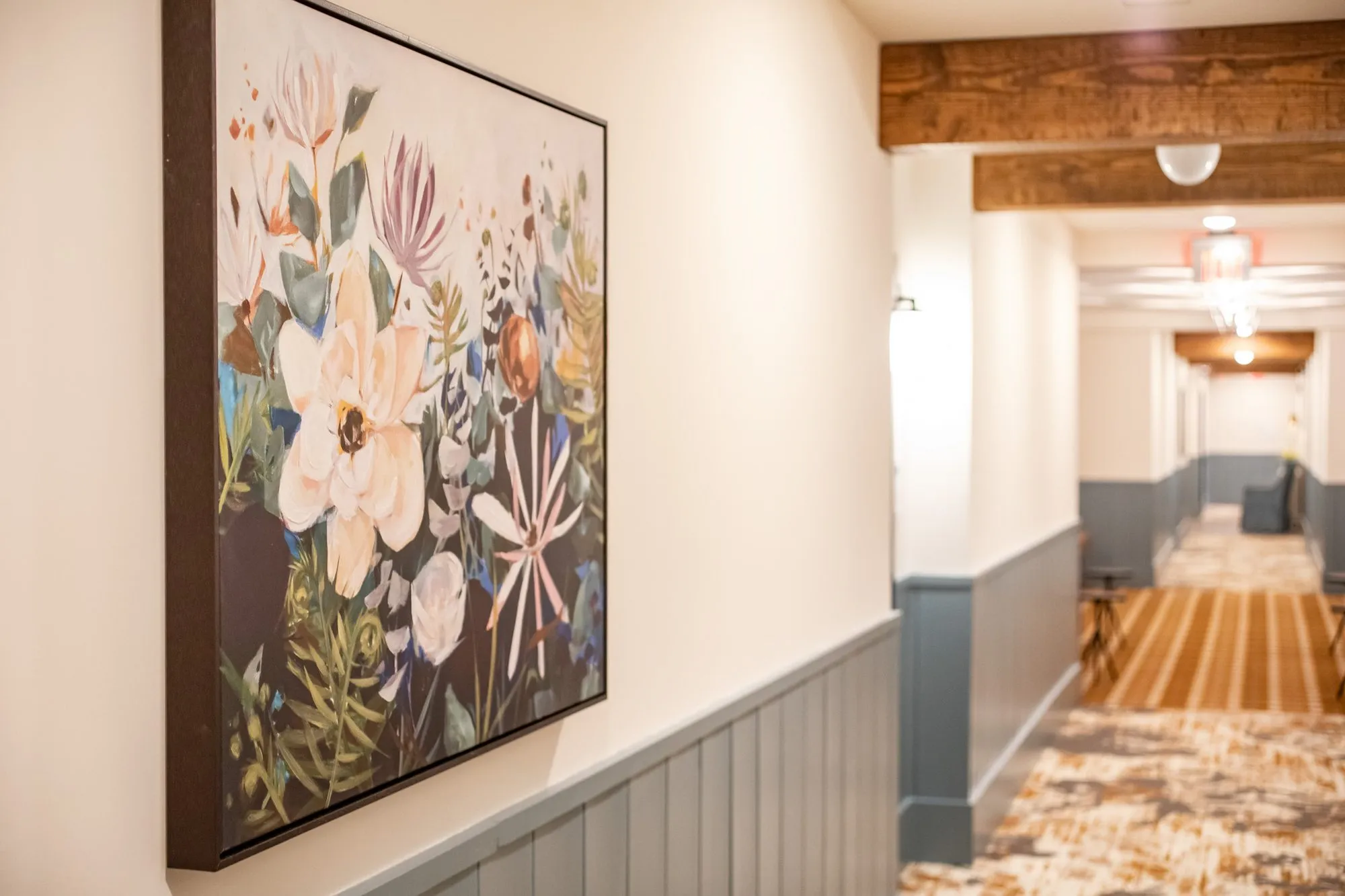 Senior living corridor with a floral painting above blue wainscot, timber beams and a plaid runner beyond