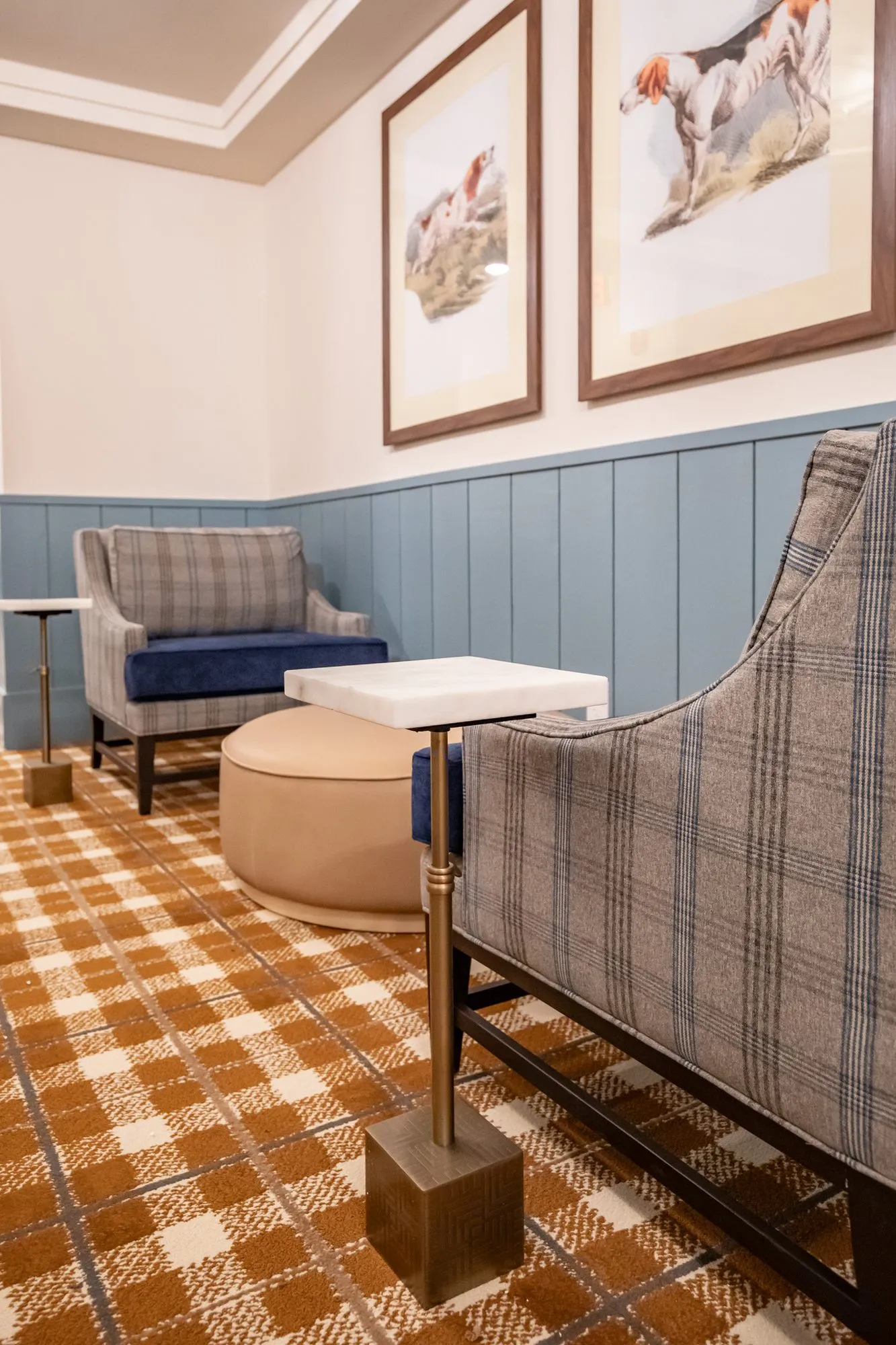 Senior living lounge with plaid armchairs, a leather ottoman and a stone topped drink table below two hunting dog prints