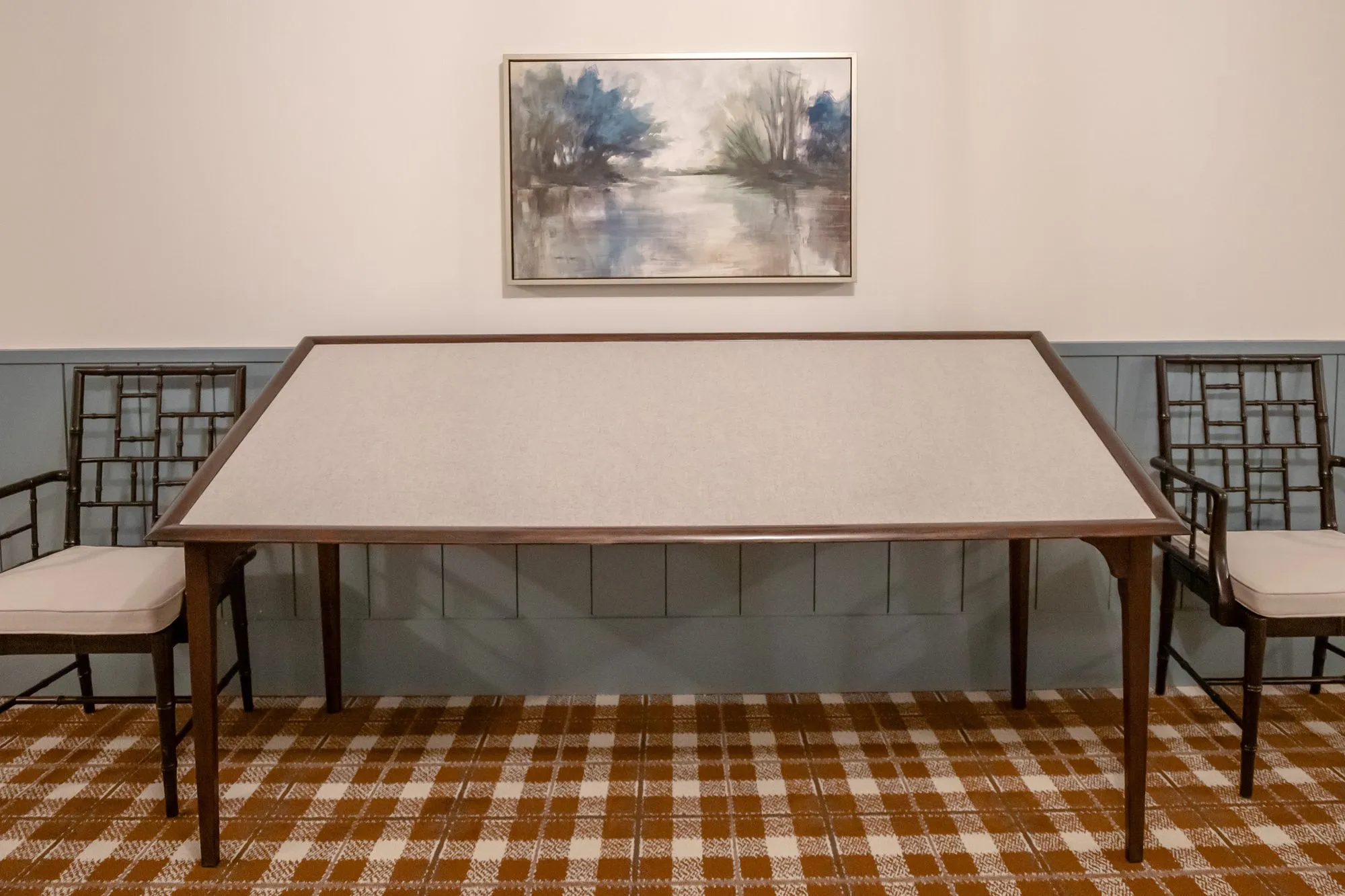 Senior living interior detail: long table with a pale top and two lattice back chairs below a lakeside painting