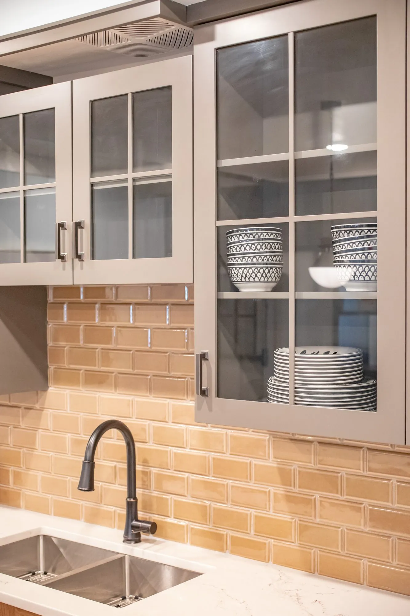 Senior living kitchenette with glass front grey cabinets holding blue and white dishes above tan subway tile