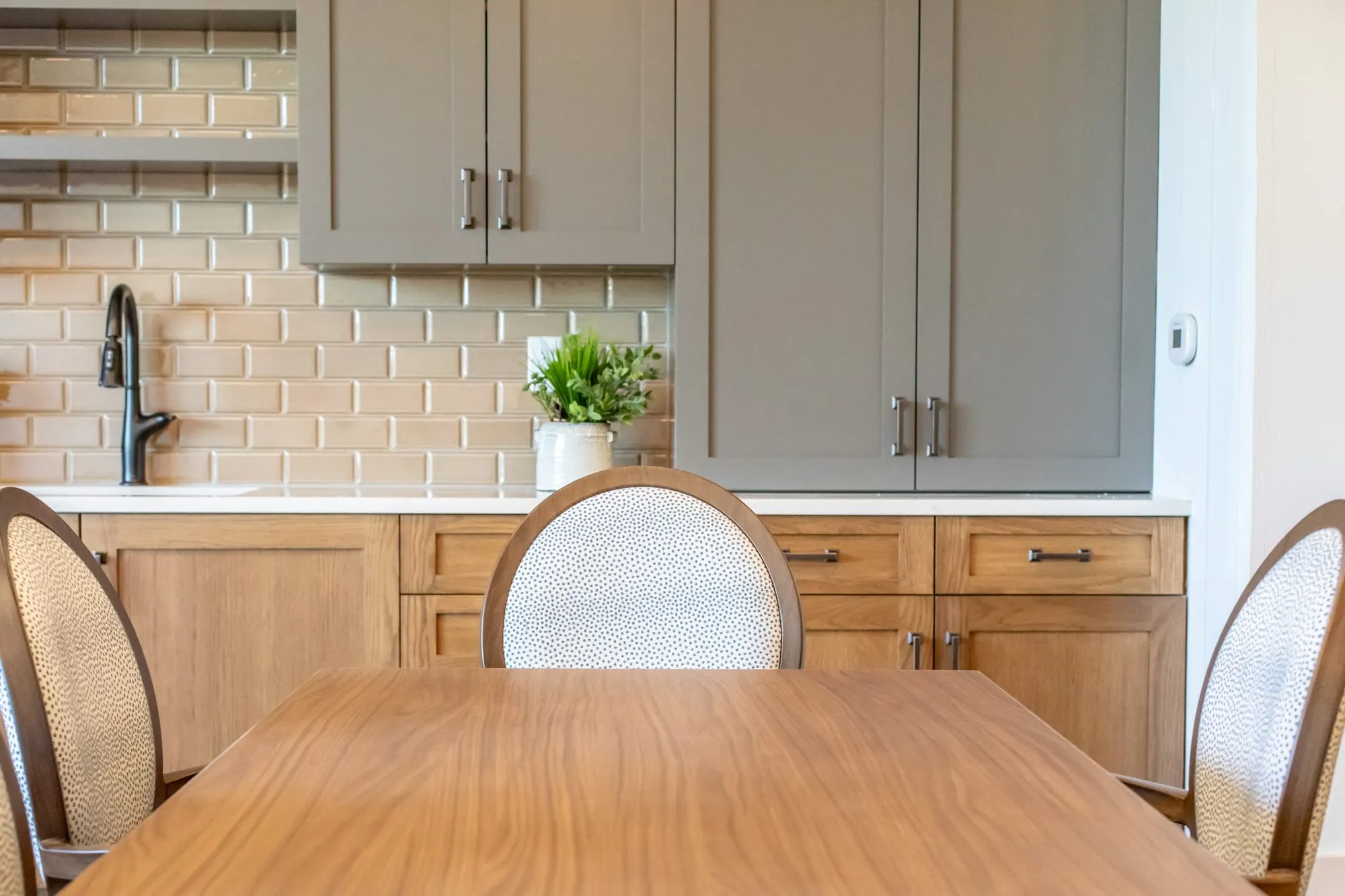 Senior living dining table with dotted fabric oval back chairs before oak and grey cabinets with tan subway tile