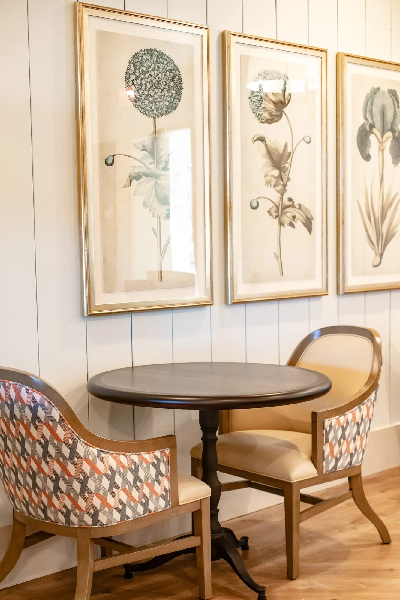 Senior living dining area with a bistro table and two geometric pattern armchairs below three botanical prints on shiplap
