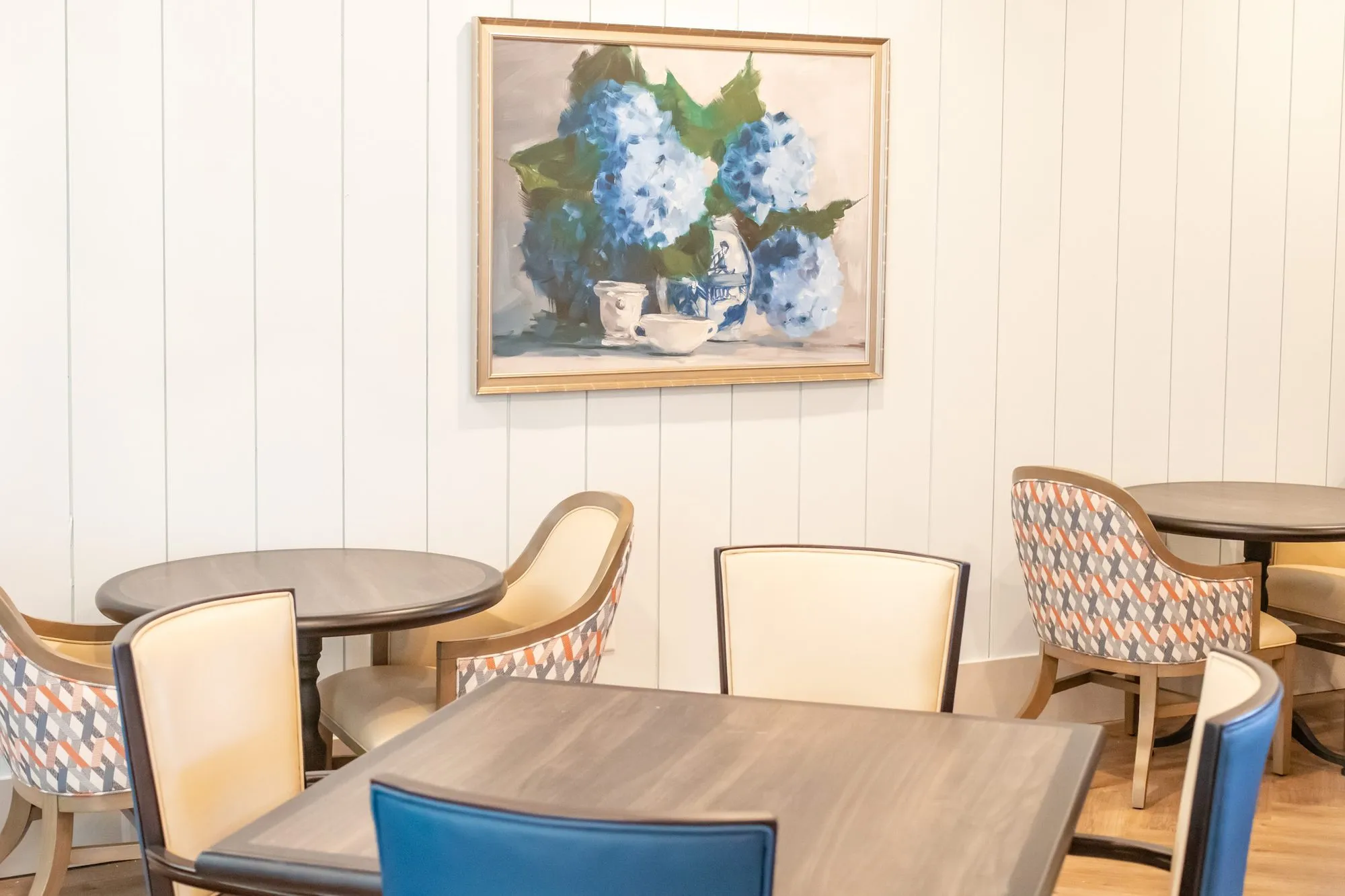 Senior living dining room with cream and blue leather chairs and geometric armchairs at tables below a hydrangea still life