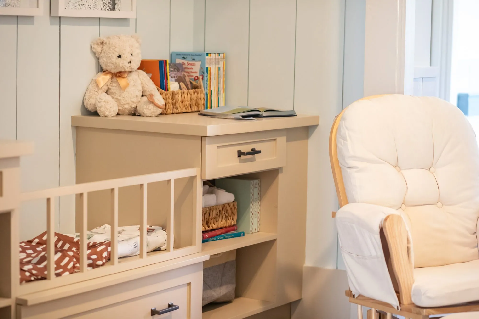 Senior living nursery corner with a cream glider, a crib, and a teddy bear and books on the dresser