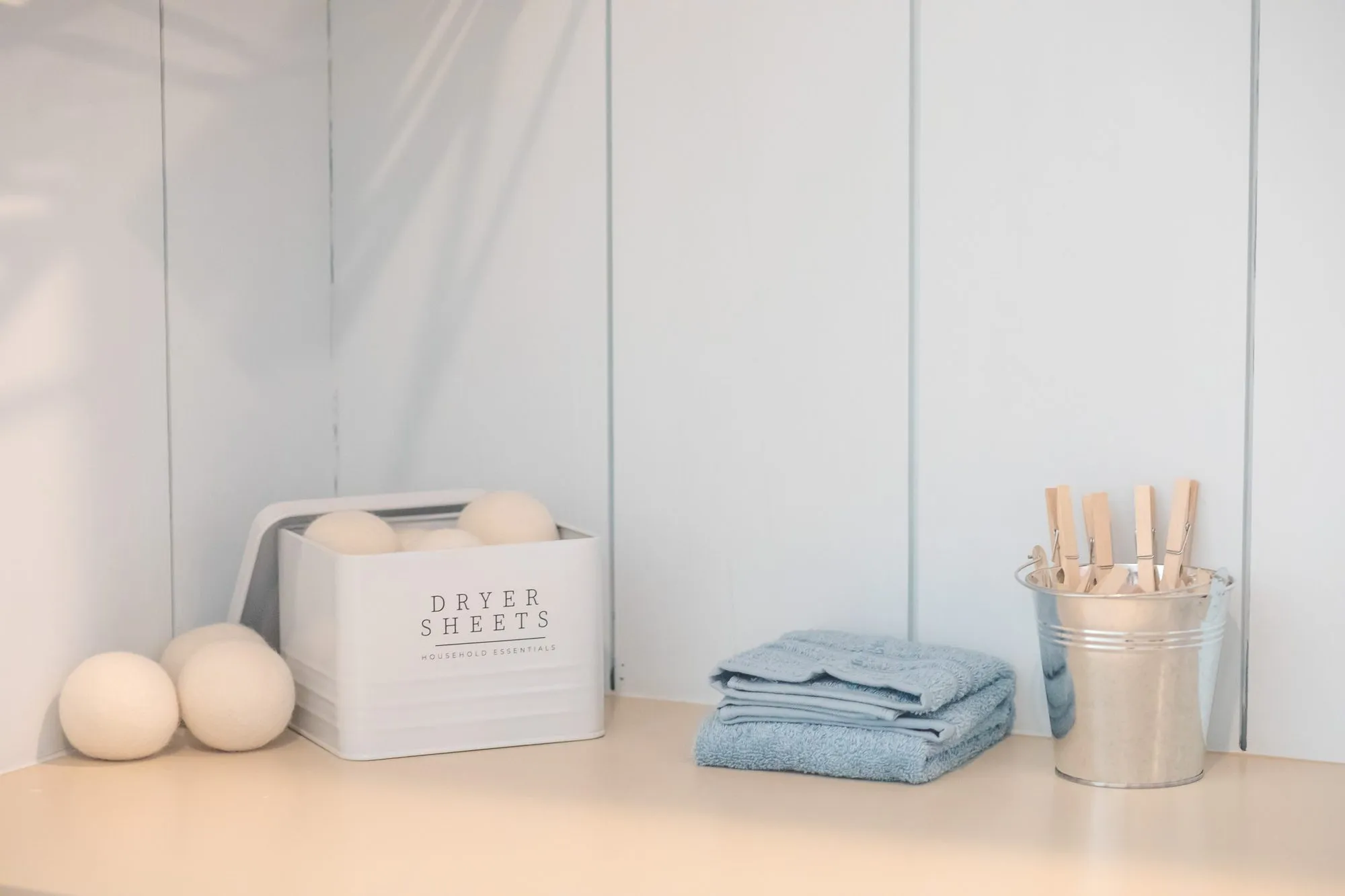 Senior living laundry counter with a dryer sheet tin, wool dryer balls, folded blue towels and a pail of clothespins