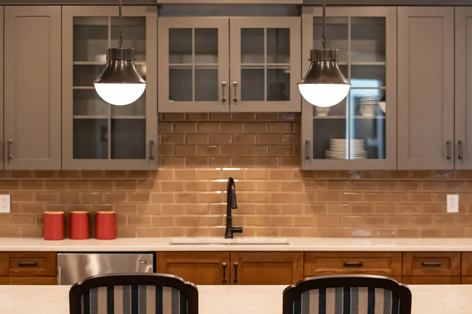 Senior living kitchenette with grey glass front cabinets, bronze pendants, tan subway tile and red canisters