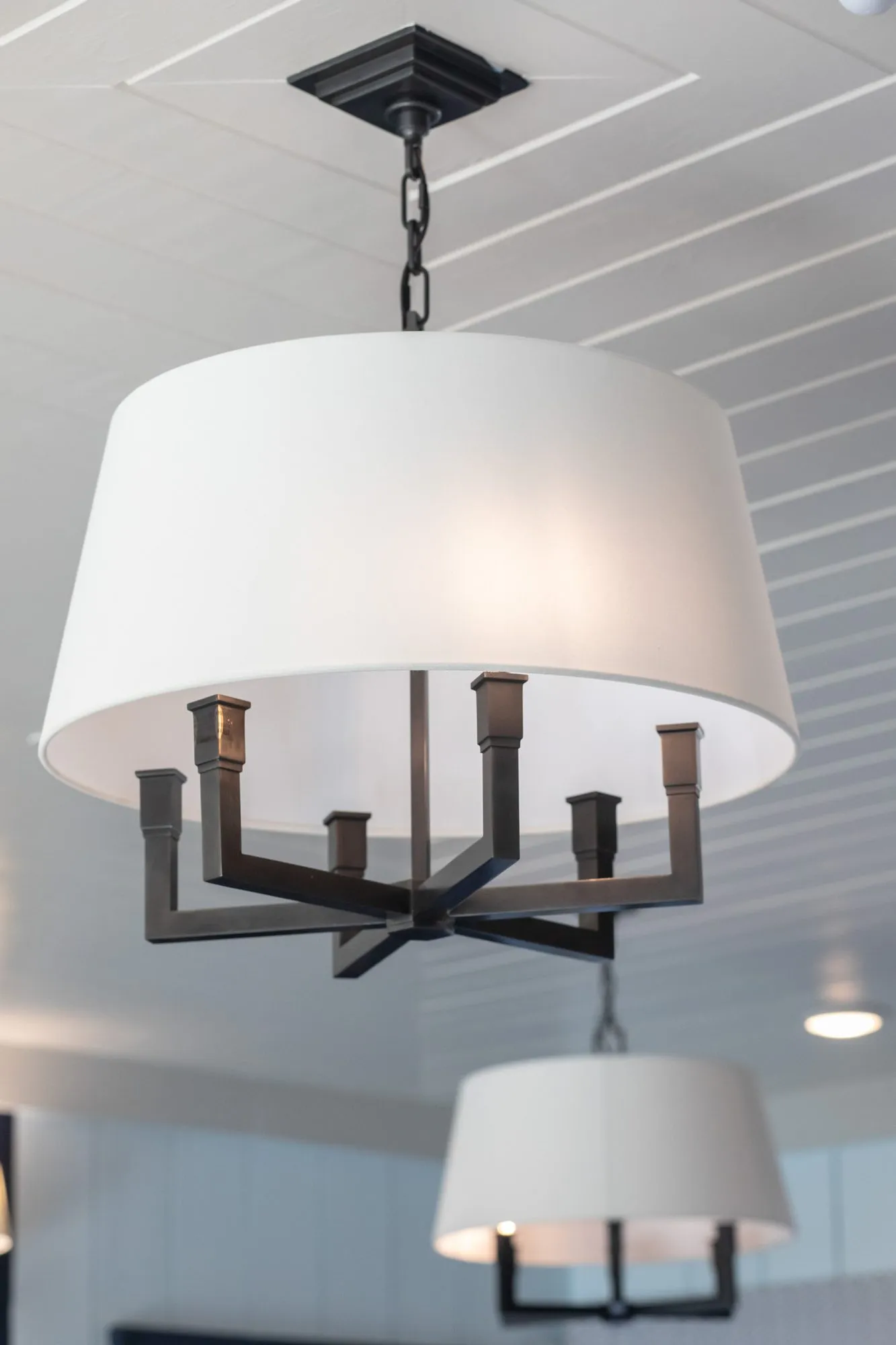 Senior living interior detail: looking up at two white drum shade chandeliers with black arms below a white plank ceiling