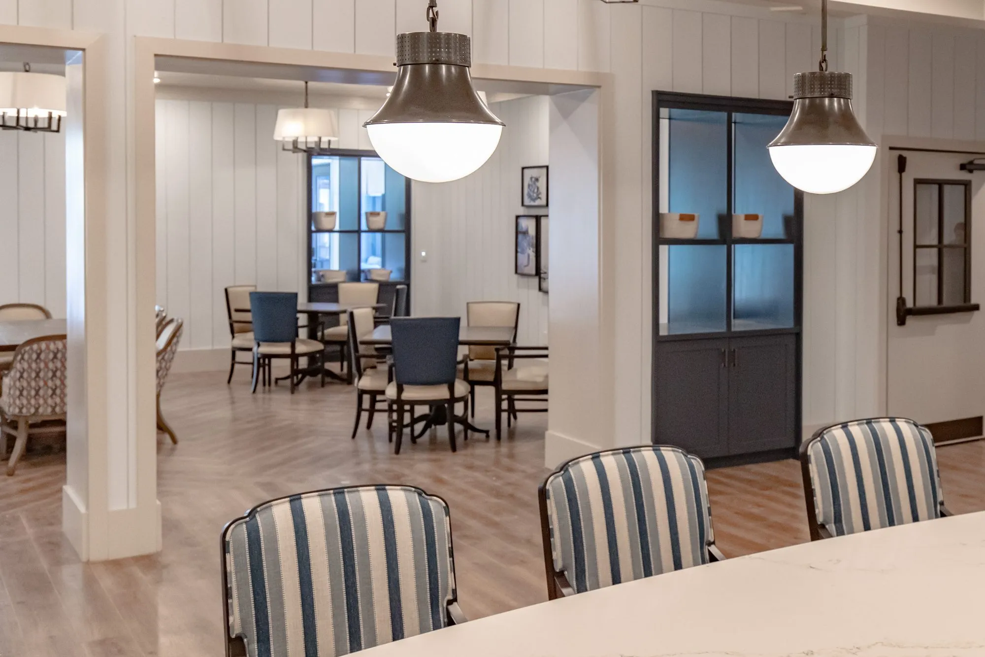 Senior living dining area through square columns, blue striped chairs at a counter in front under bronze pendants