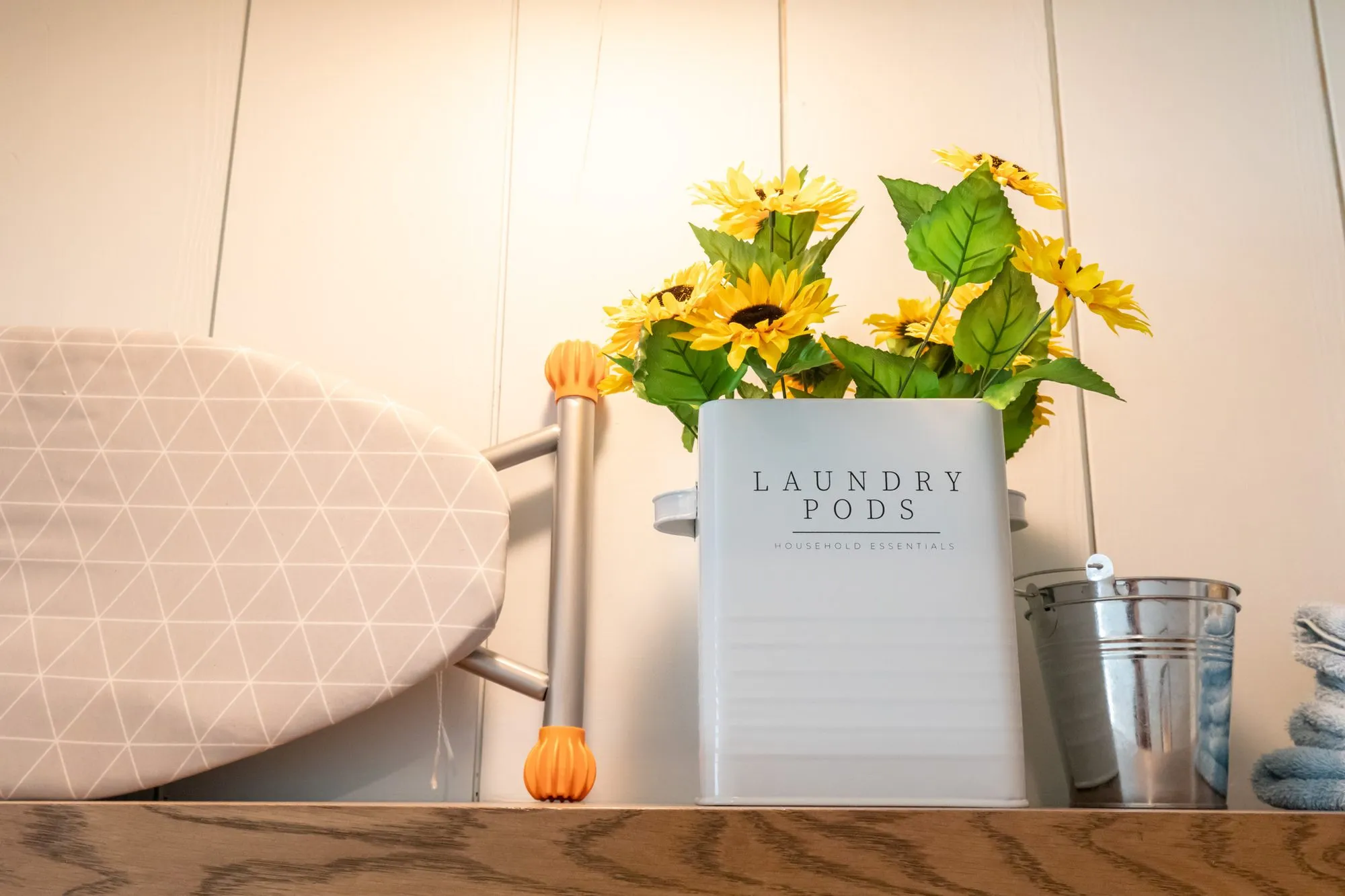 Senior living interior detail: ironing board, a laundry pod canister with sunflowers and a galvanised pail on a shelf