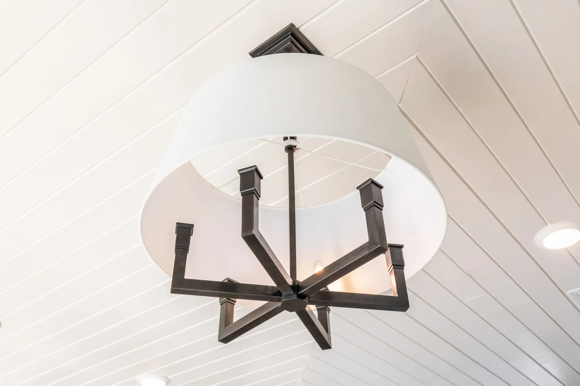 Senior living interior detail: looking up at a white drum shade chandelier with black arms on a herringbone plank ceiling