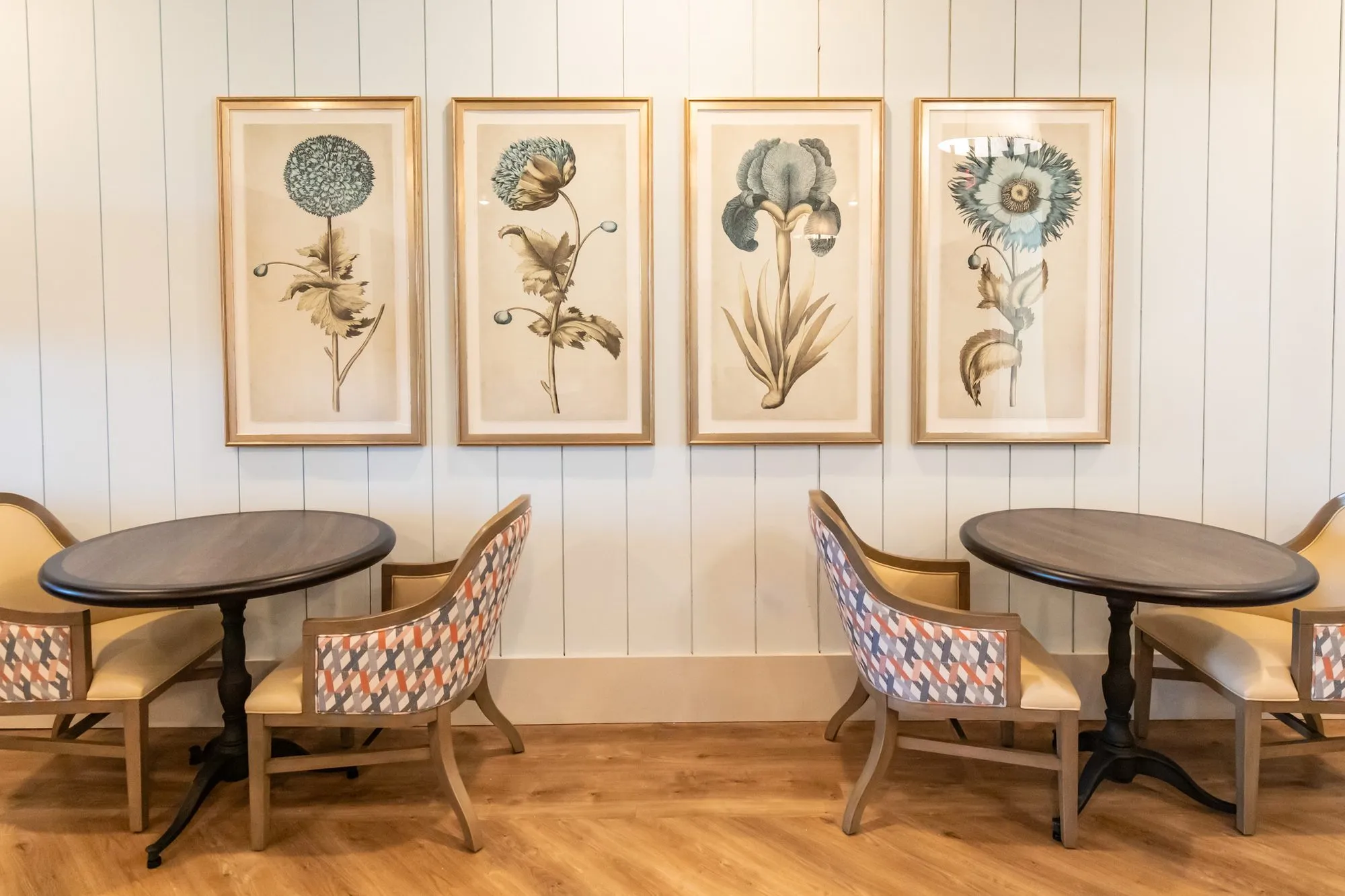 Senior living dining area with two bistro tables and geometric pattern chairs below four framed botanical prints on shiplap