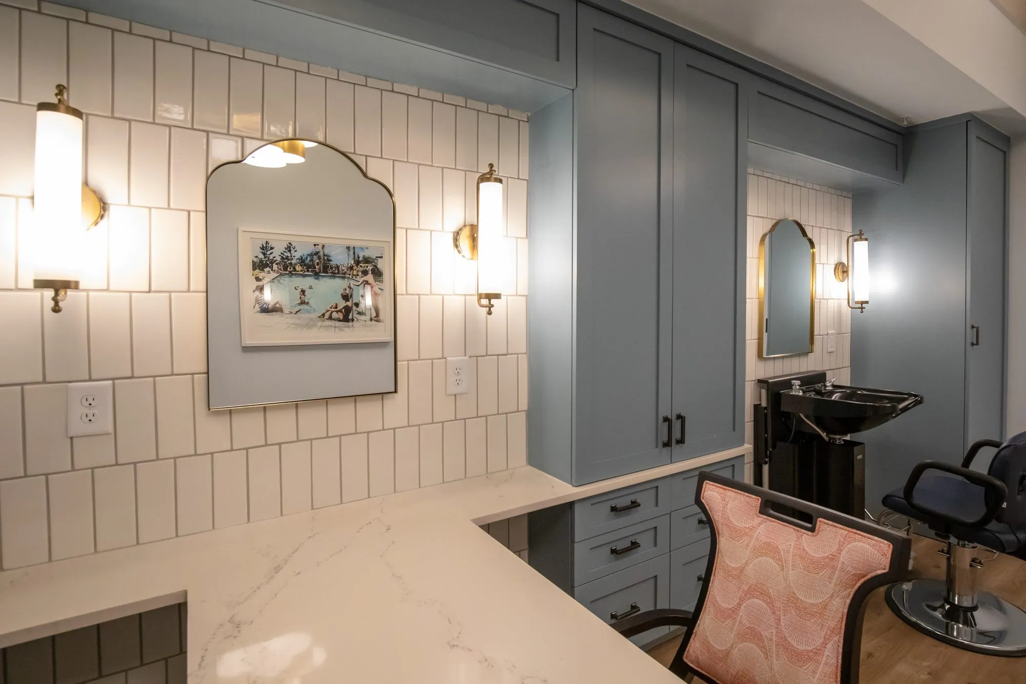 Senior living salon with blue cabinetry, arched brass mirrors and glass sconces on white stacked tile, a shampoo chair