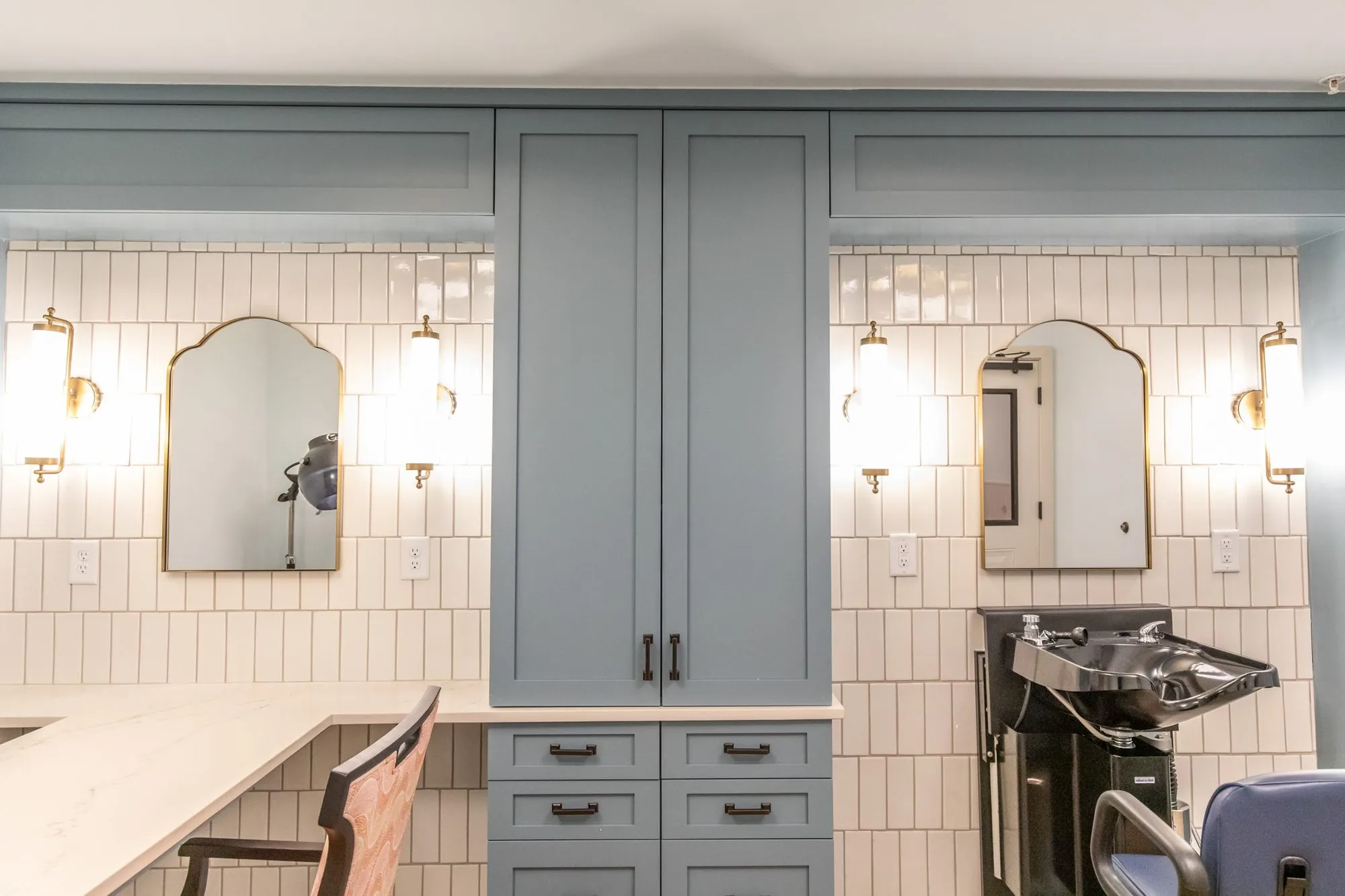 Senior living salon wall of blue cabinets between two arched brass mirrors with sconces, a shampoo bowl at right