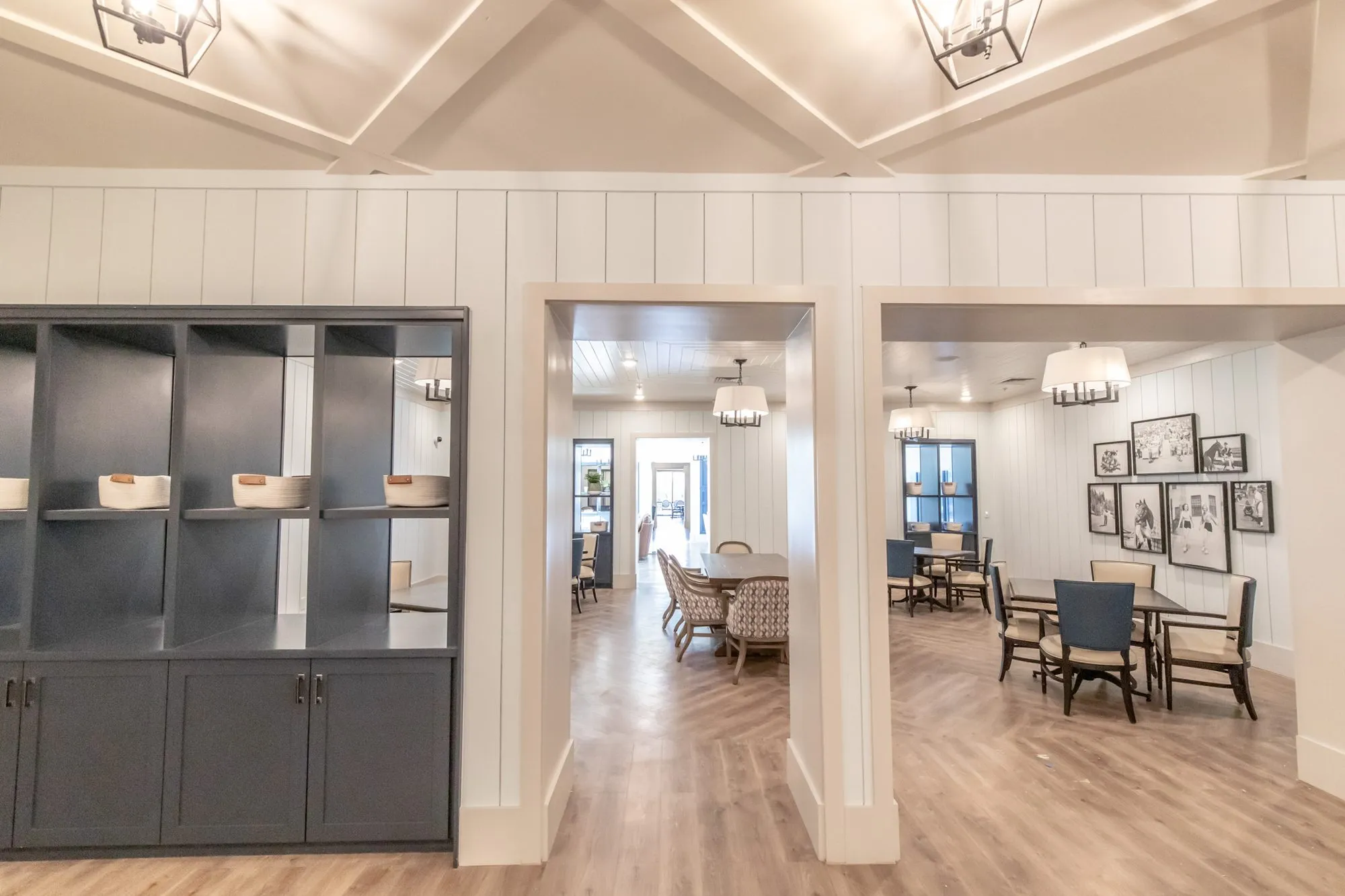 Senior living hall with navy open shelving beside wide doorways into a dining room with framed photos, lantern pendants above