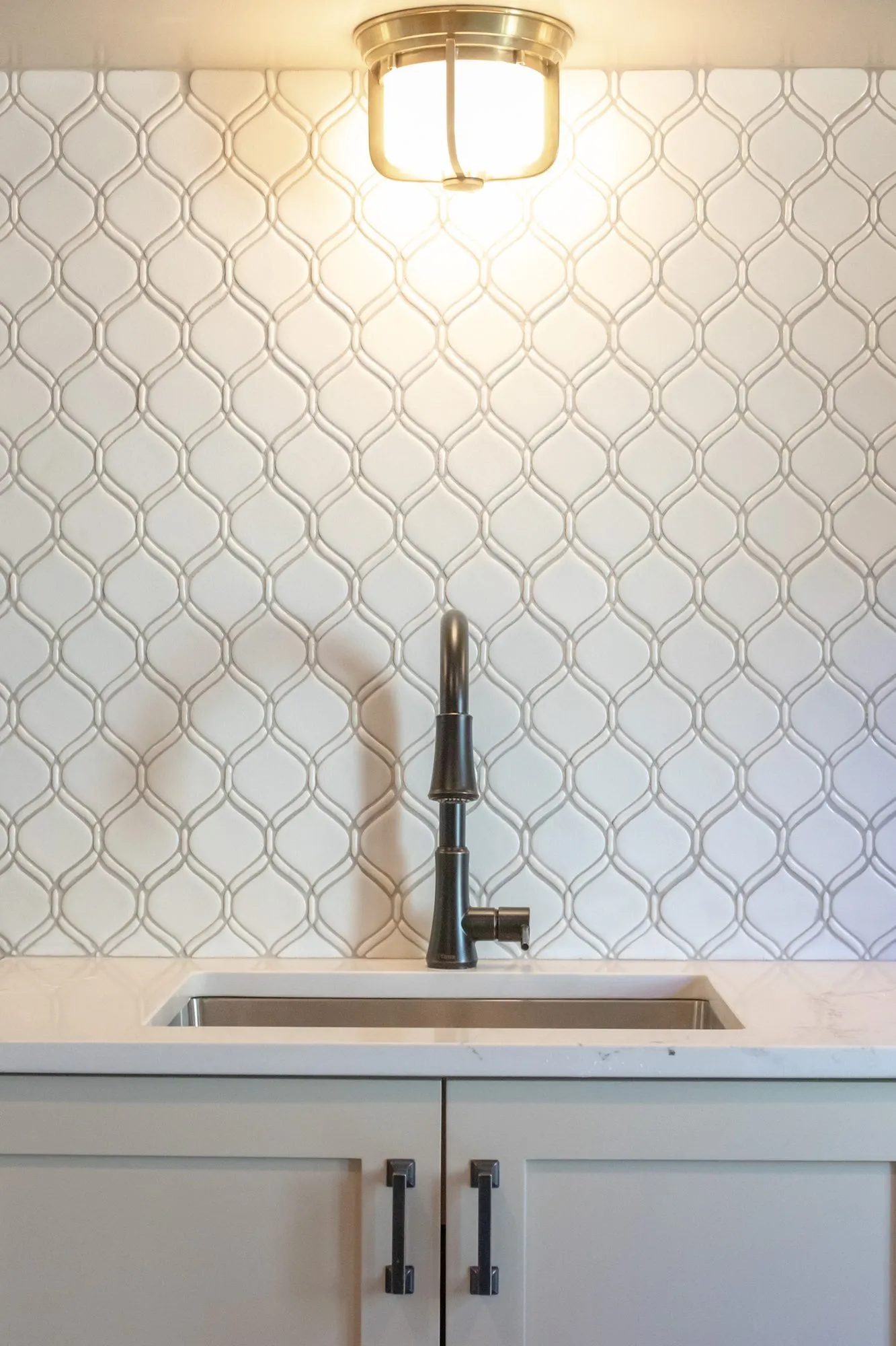 Senior living interior detail: black faucet over a stainless sink below a brass flush light on white ogee tile