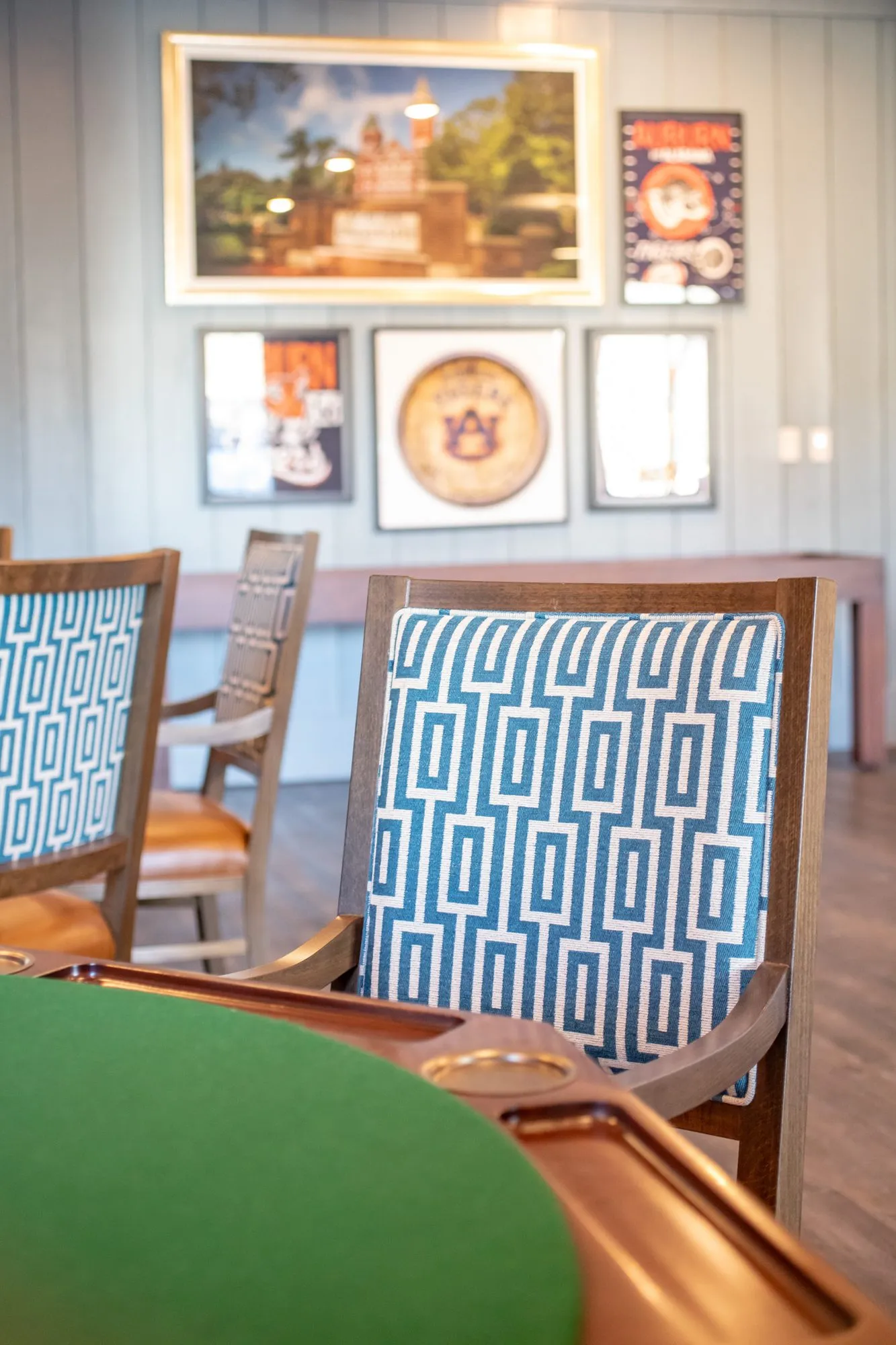 Senior living interior detail: Greek key chair at a green felt card table, college posters on the shiplap beyond