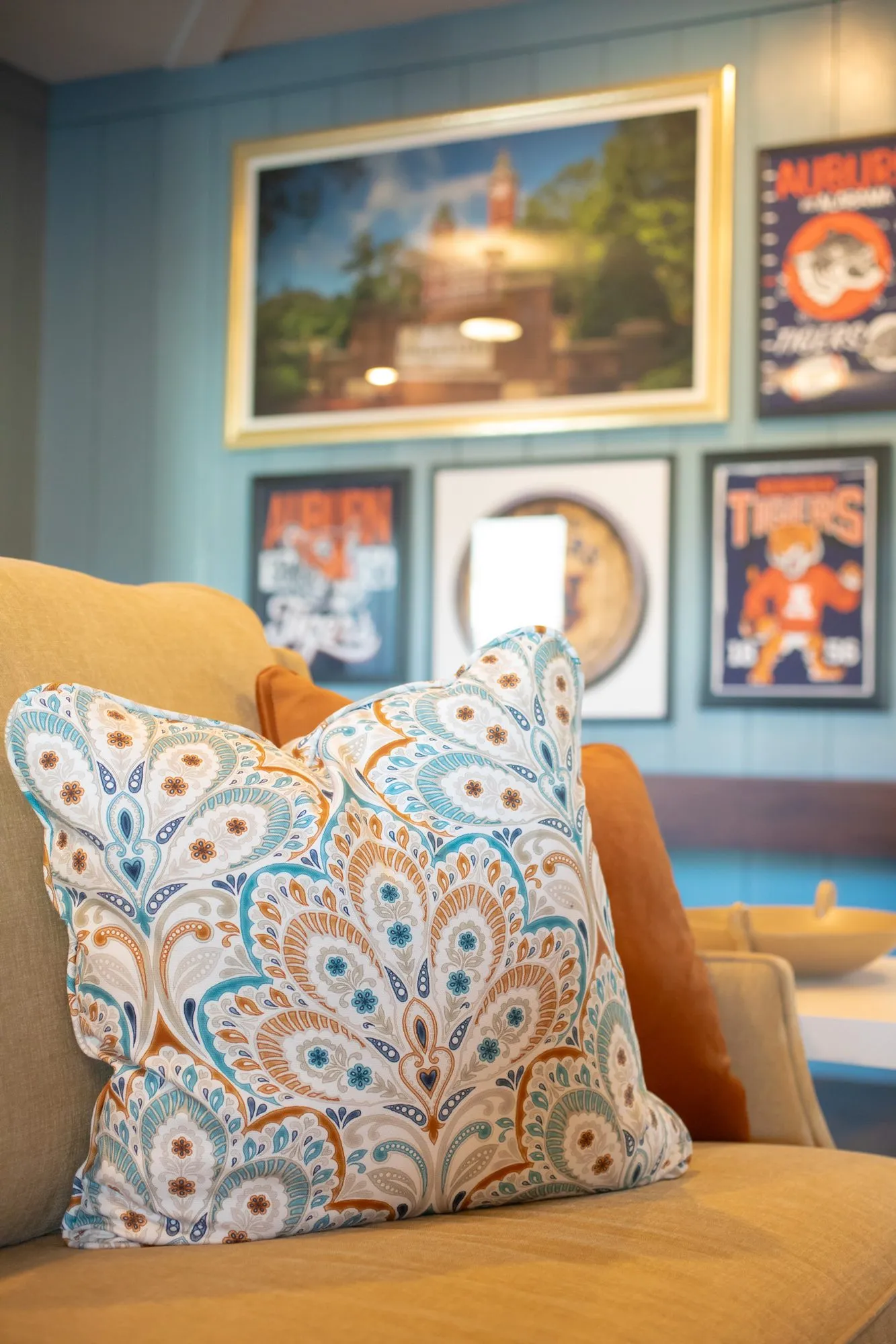 Senior living interior detail: paisley pillow on a camel sofa below Auburn posters and a campus painting