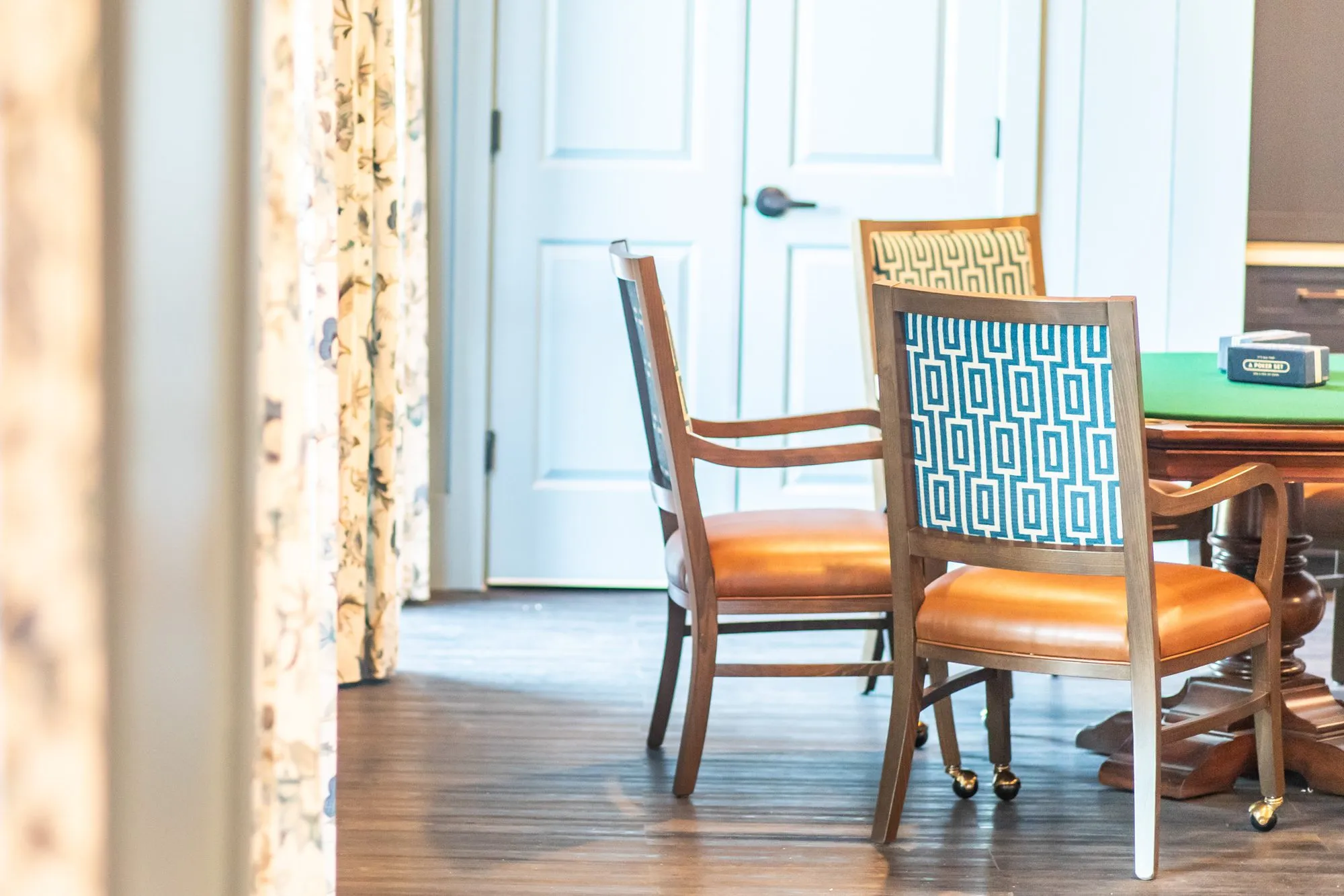 Senior living card room with a card table and leather seated Greek key chairs past floral drapery, white double doors behind