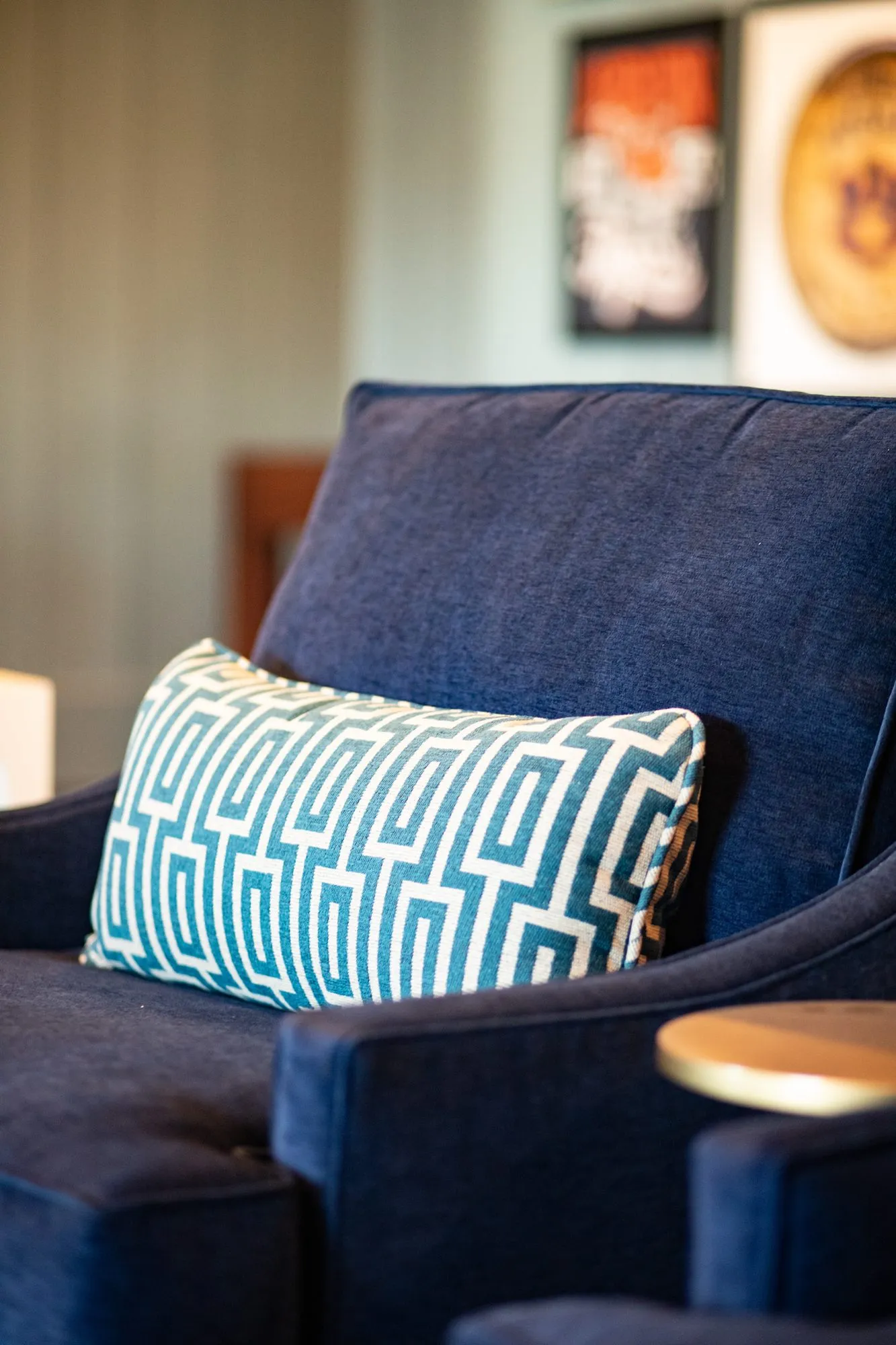 Senior living interior detail: navy velvet armchair with a teal Greek key pillow, framed posters blurred behind