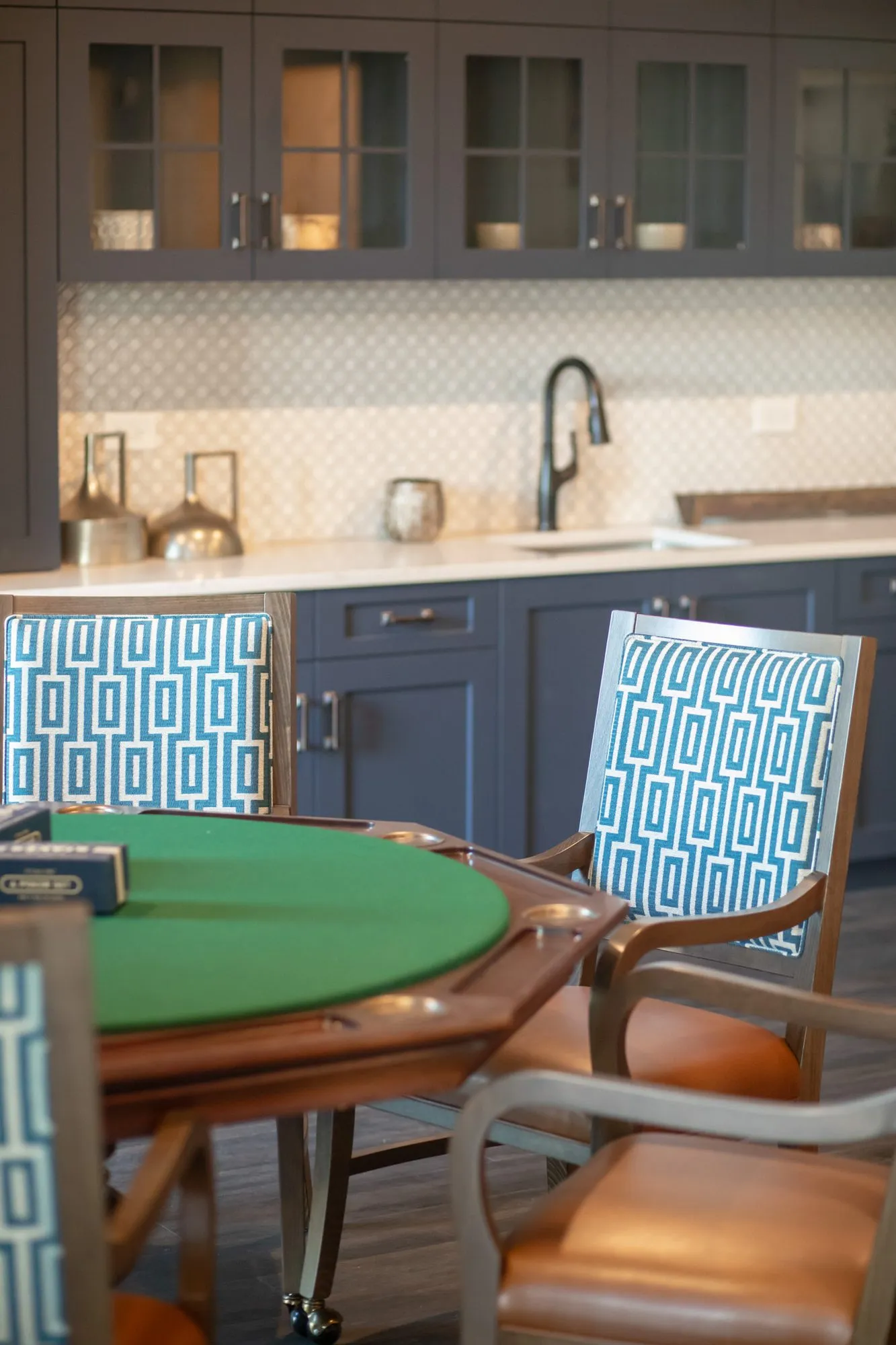 Senior living card room with a green felt card table and Greek key chairs, navy kitchenette cabinets behind
