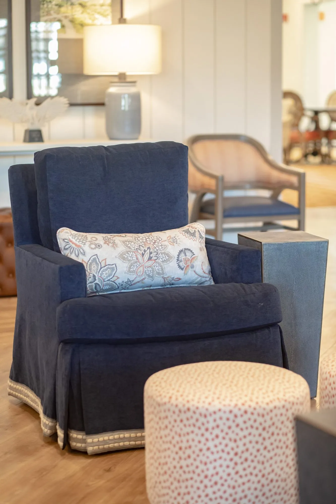 Senior living interior detail: navy skirted armchair with a floral pillow and a dotted pouf