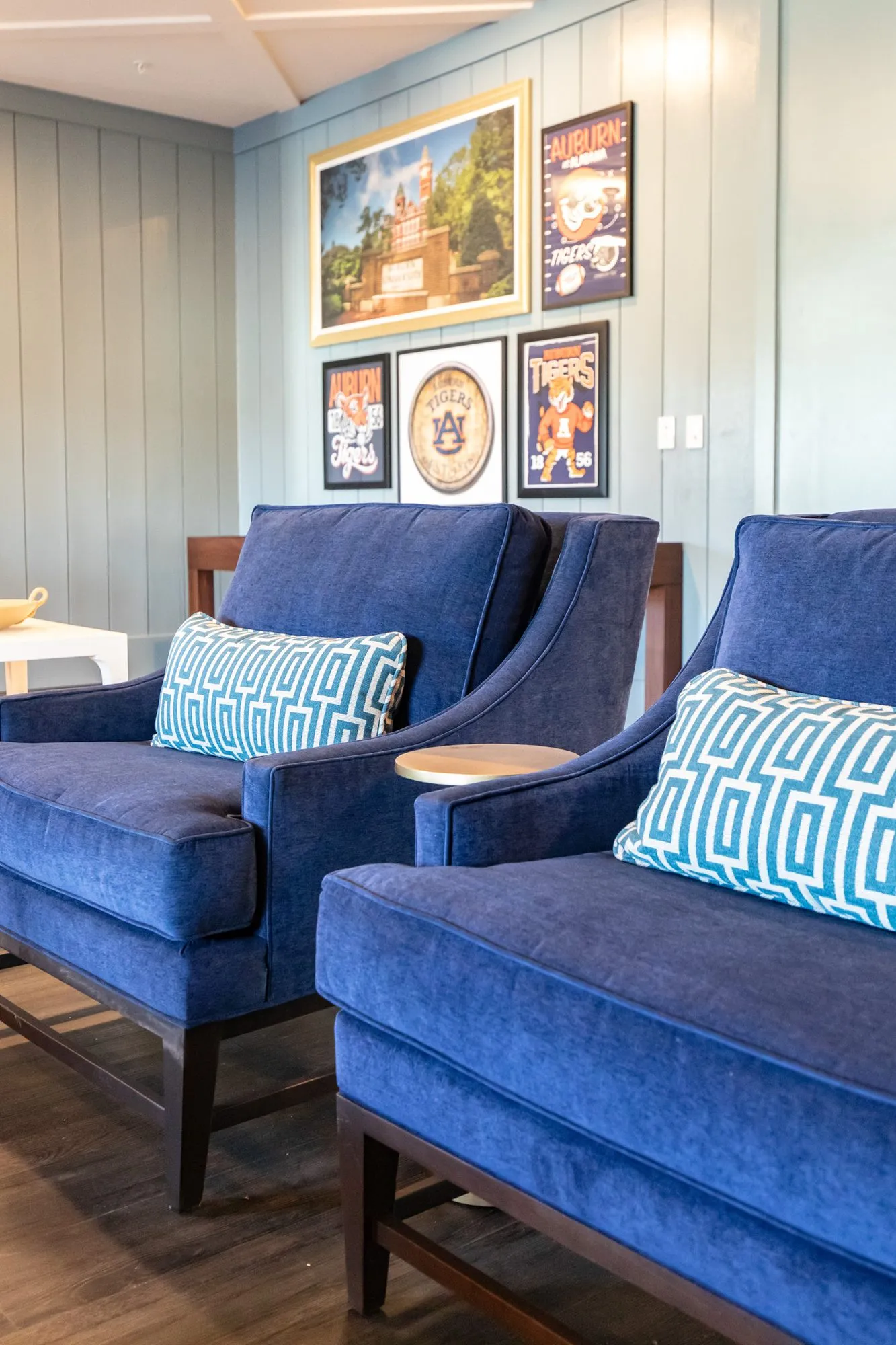 Senior living interior detail: two navy velvet armchairs below Auburn posters and a campus painting on blue shiplap