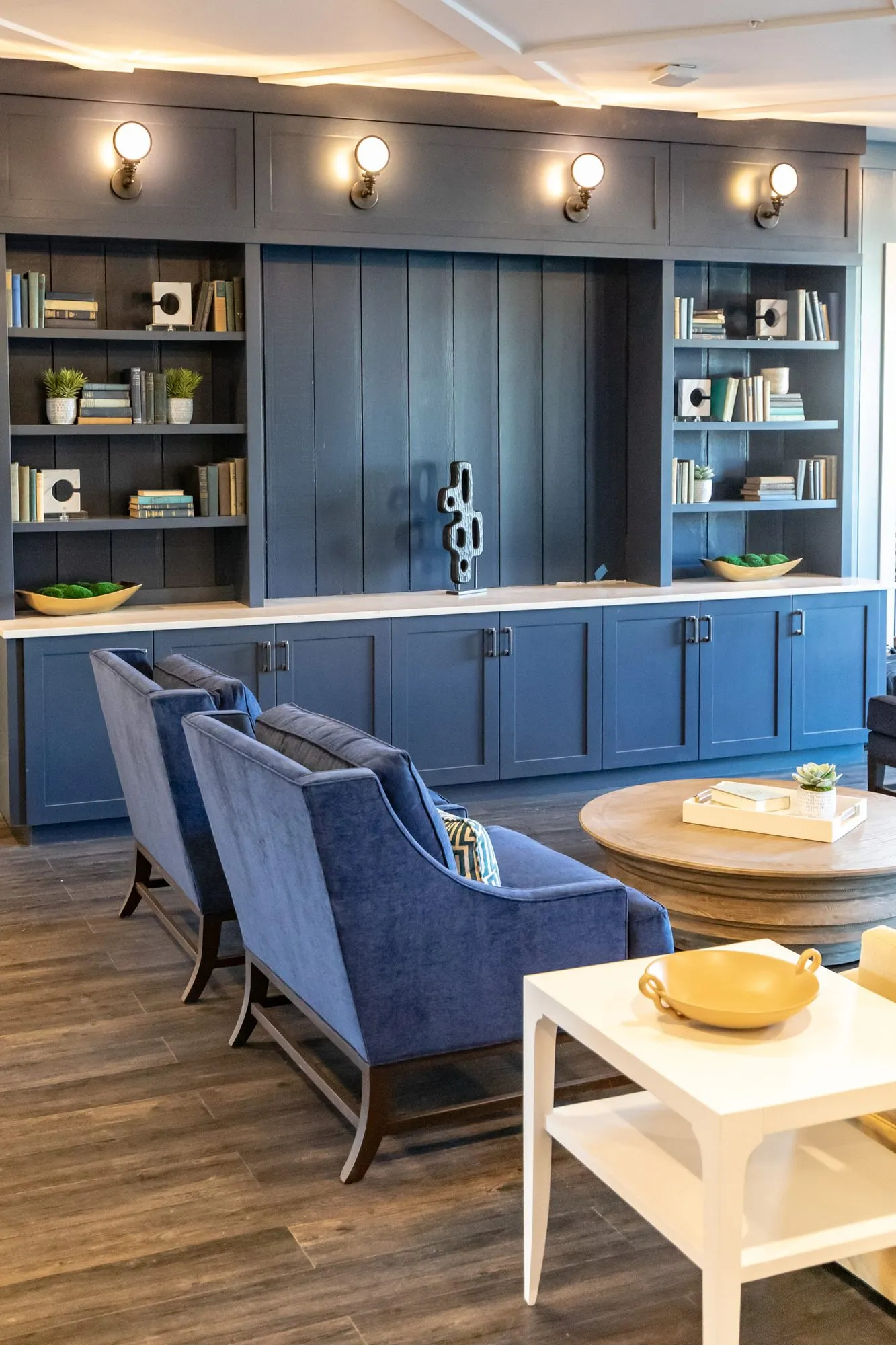 Senior living lounge with navy armchairs and a round coffee table before navy built-ins with sconces