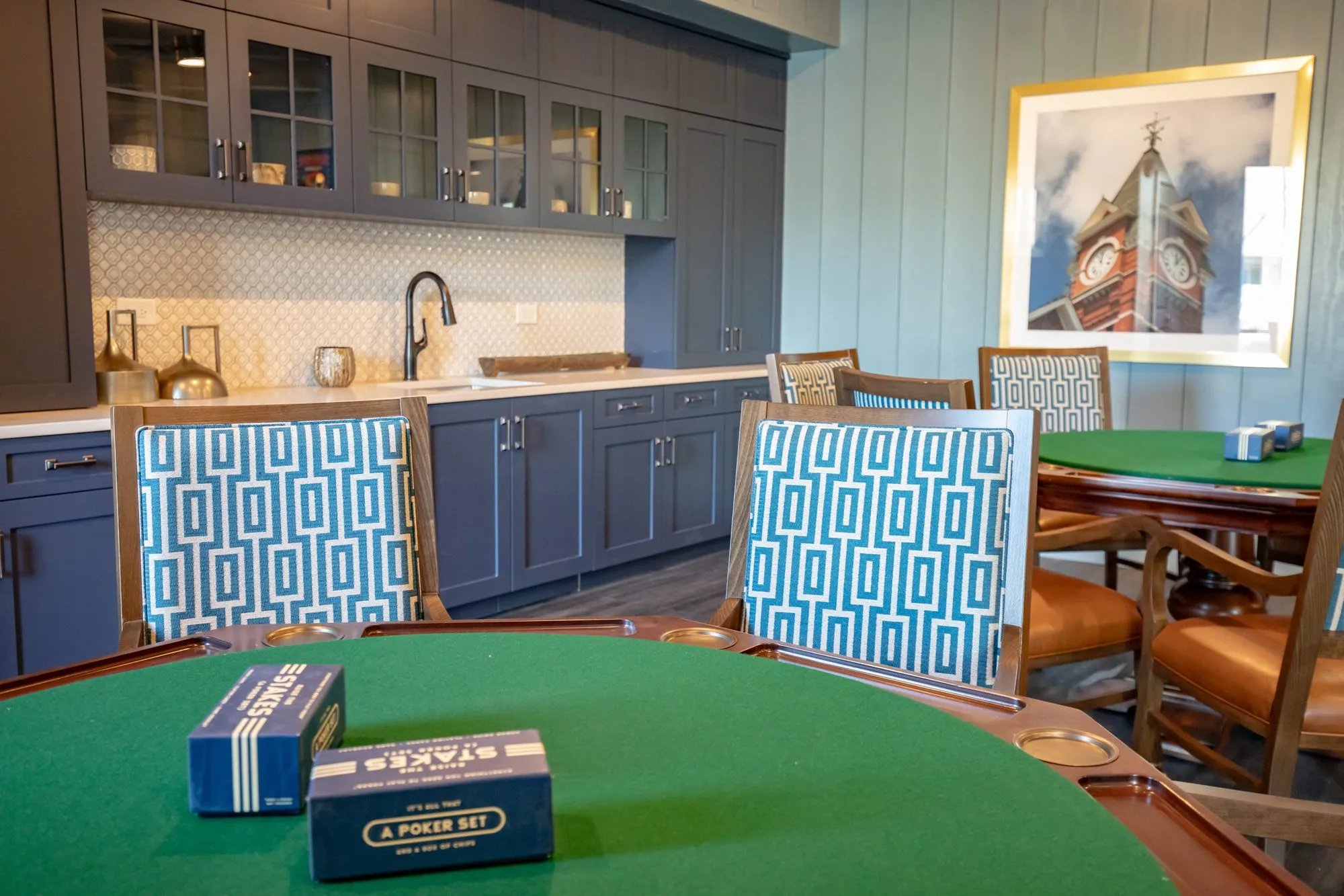 Senior living card room with boxed poker sets on a green felt table, Greek key chairs and a clock tower photograph beyond
