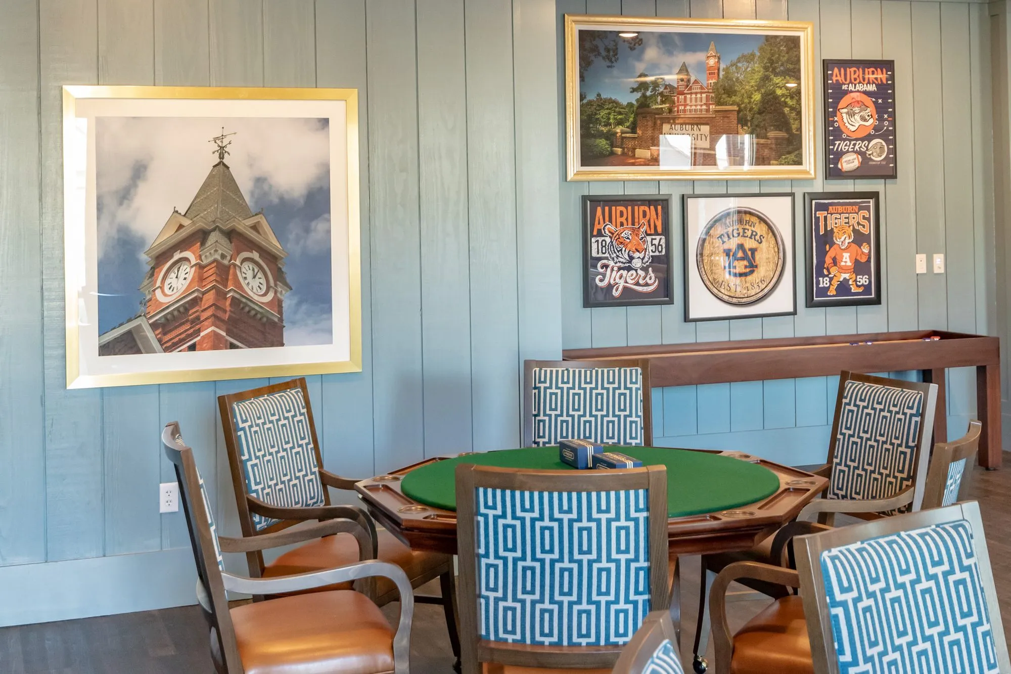 Senior living card room with a card table and Greek key chairs below a clock tower photograph and Auburn posters on shiplap