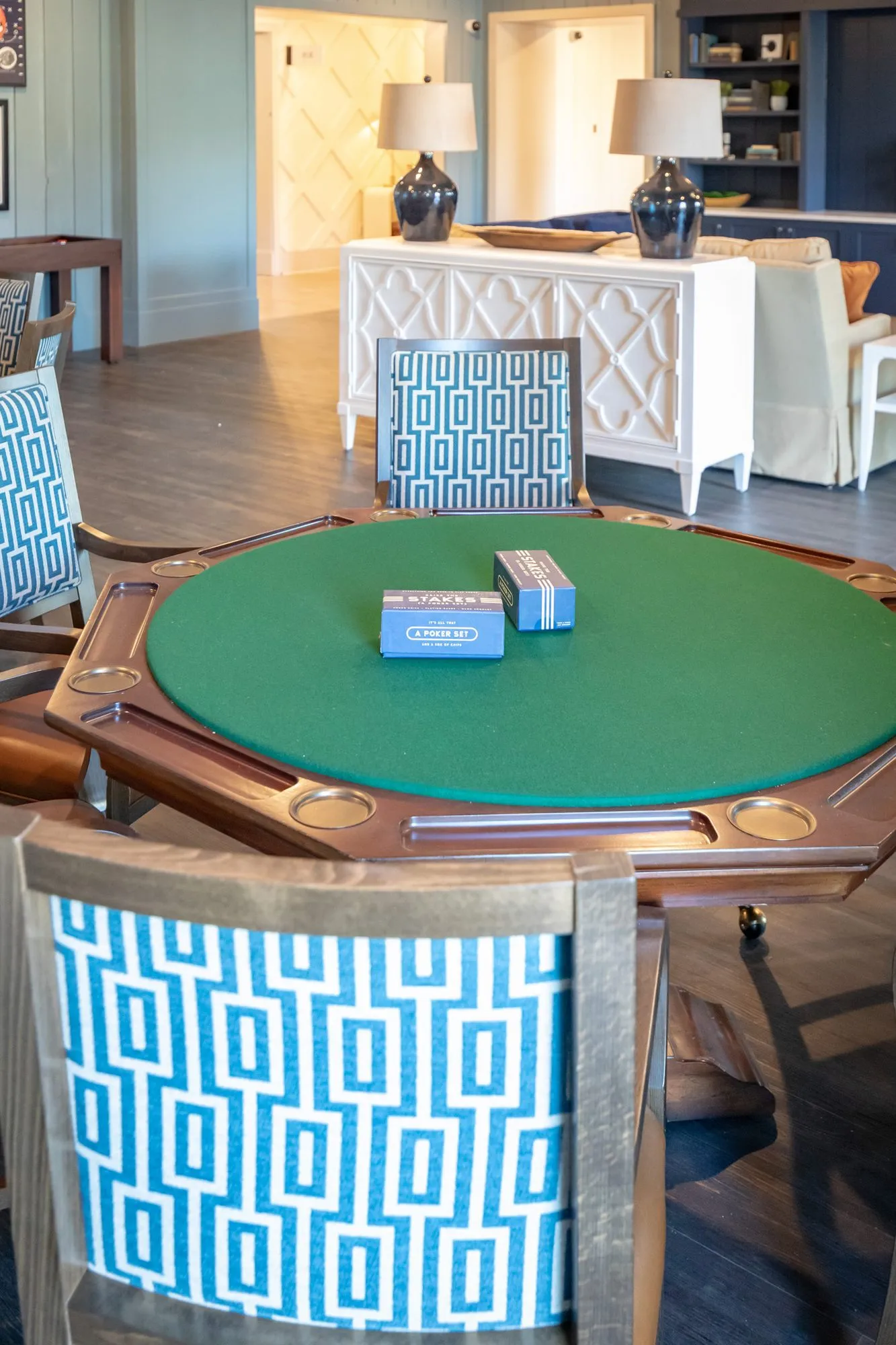 Senior living card room with a green felt card table and poker sets, a white console and navy lamps beyond