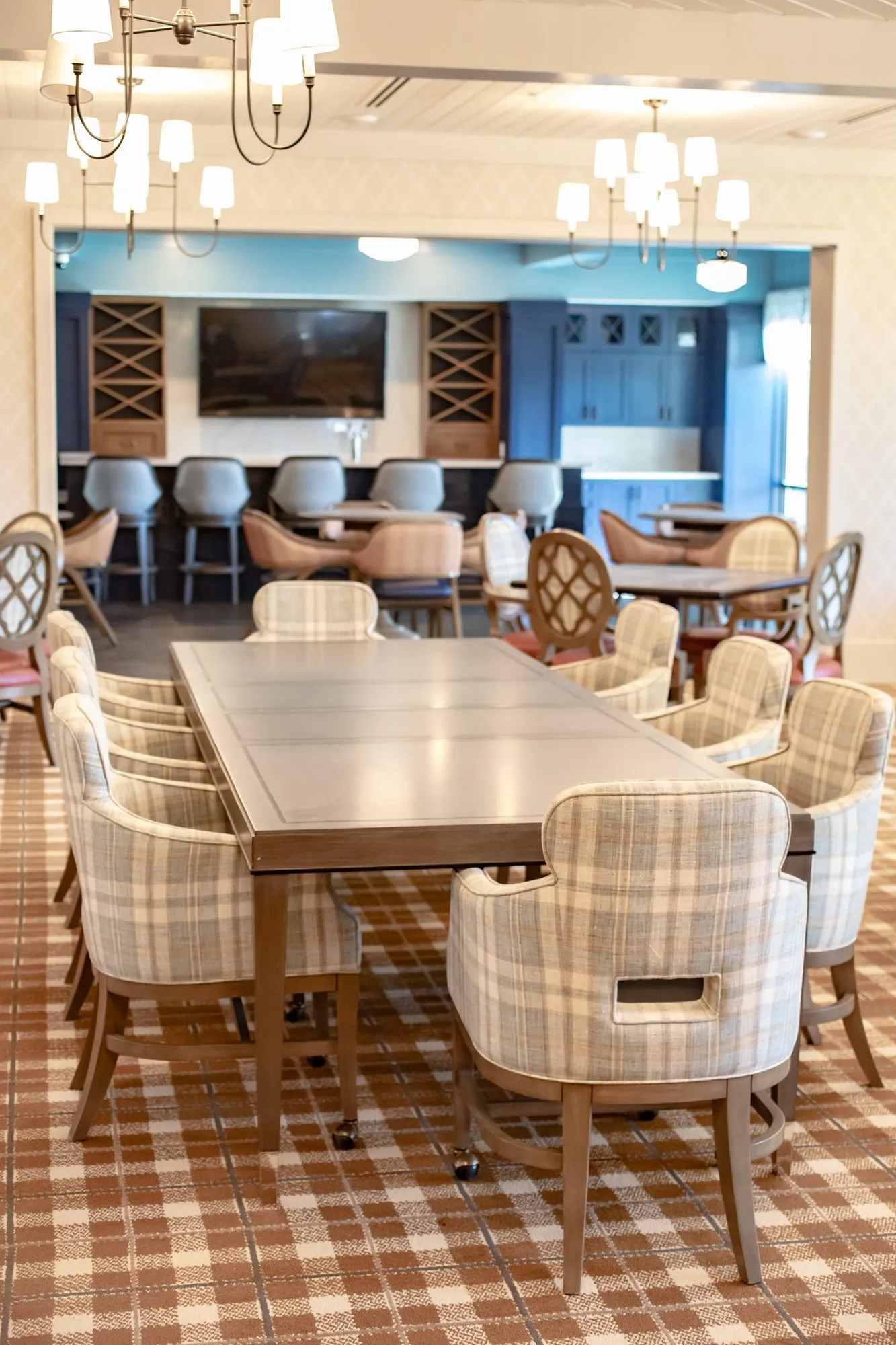 Senior living dining room with a long table of plaid chairs, a bar with barstools and a TV at the back