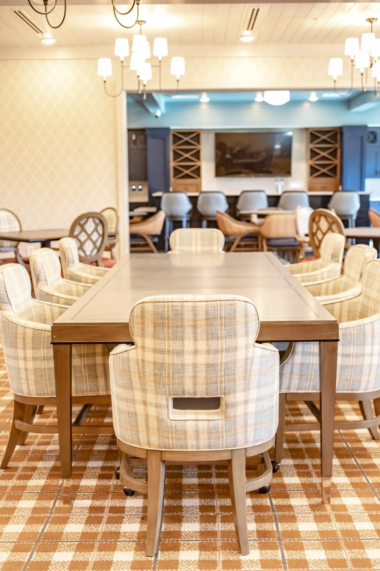Senior living dining room with a long table of plaid chairs centred toward a bar wall with a TV and wine racks