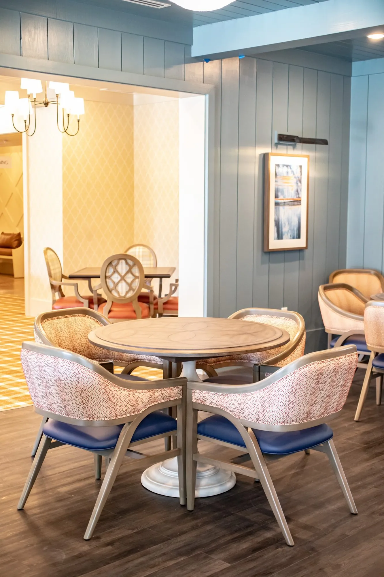 Senior living pub with coral dot chairs at a round table, blue shiplap and a doorway to the dining room