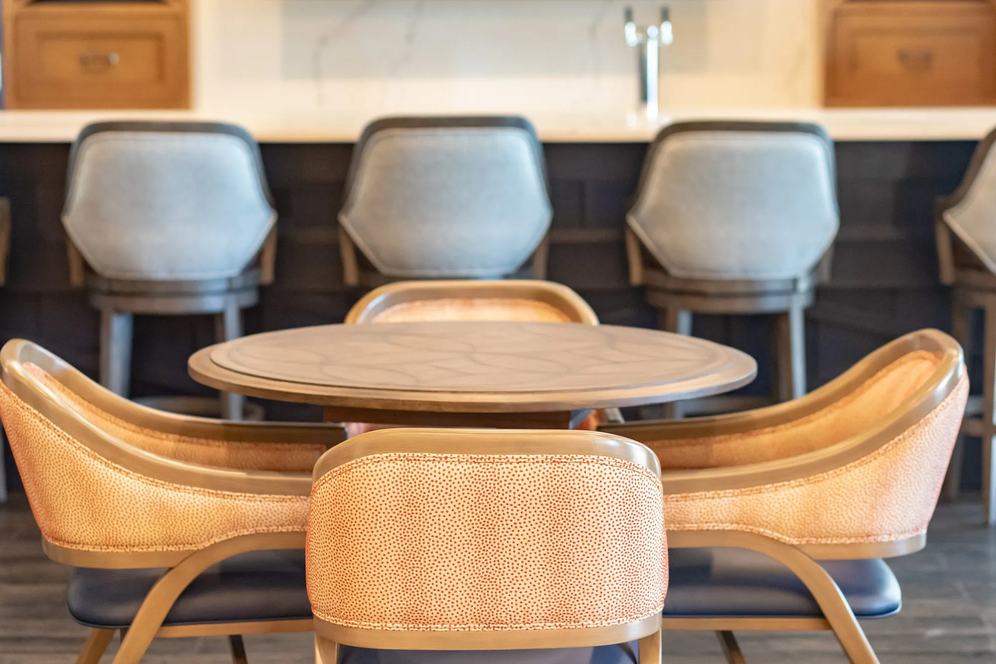 Senior living pub with four coral dot chairs at a round table before grey upholstered barstools at the bar