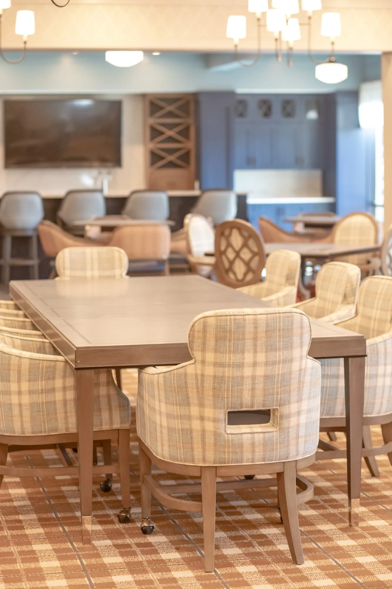 Senior living dining room with plaid chairs at long tables toward the bar and TV