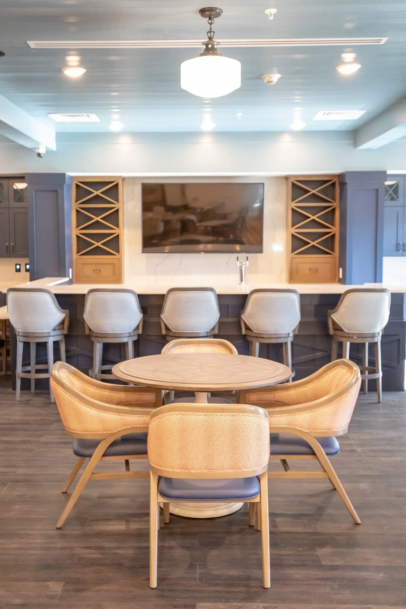 Senior living pub with a round table and coral dot chairs before the bar, TV and wine racks