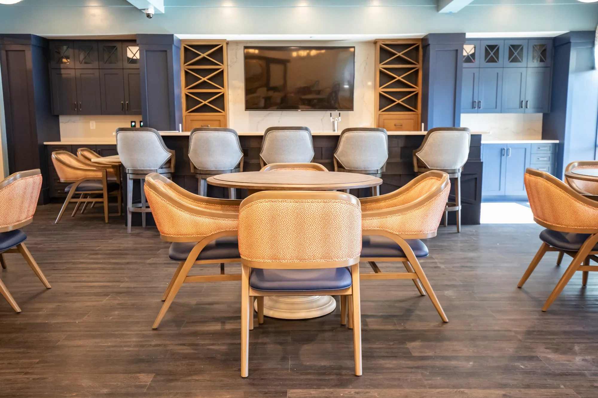 Senior living pub with a round table and coral dot chairs centred on the bar, barstools, TV and navy glass cabinets