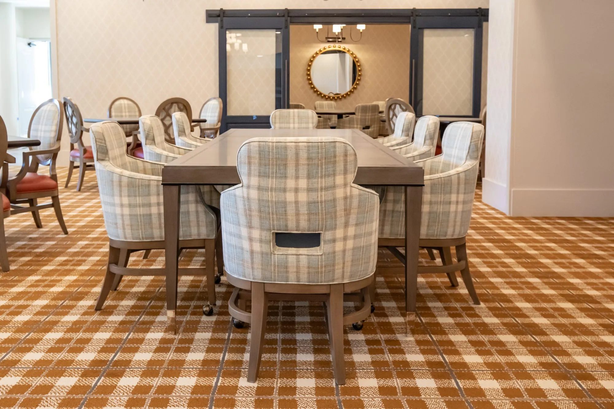 Senior living dining room with a long table of plaid chairs before barn doors to a private dining room and its gilt mirror