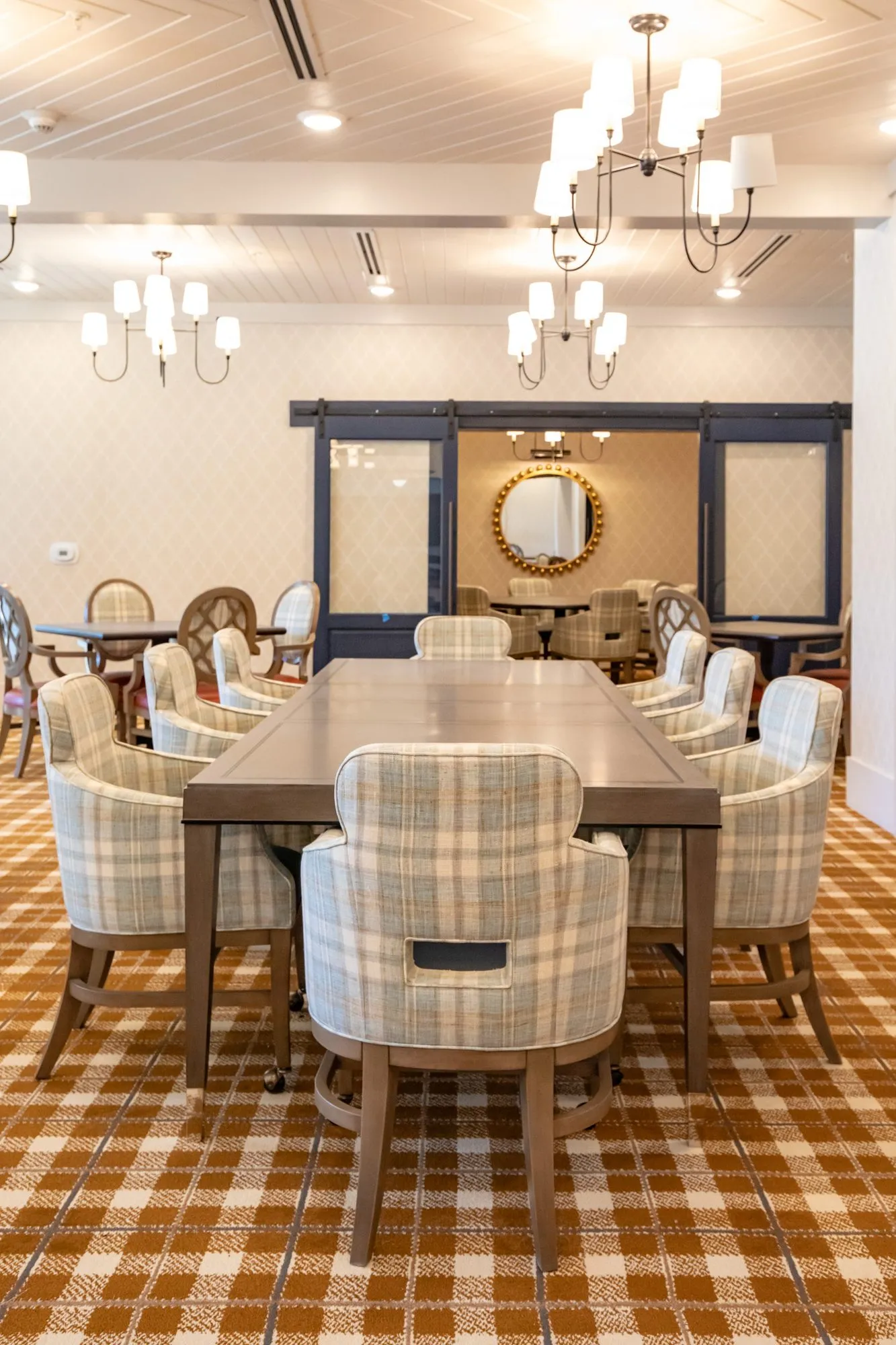 Senior living dining room with a long table of plaid chairs under candle chandeliers toward the barn doors
