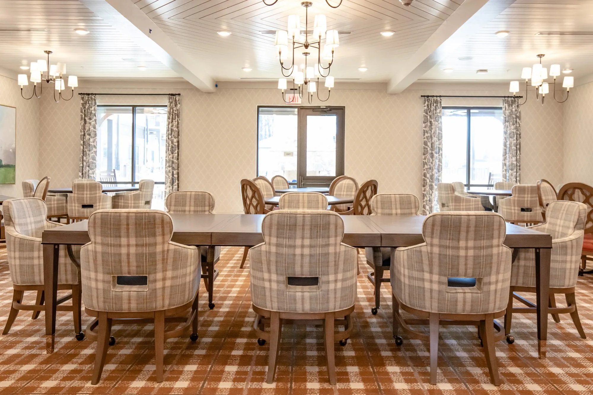 Senior living dining room with plaid chairs at long tables under candle chandeliers, curtained windows along the back wall