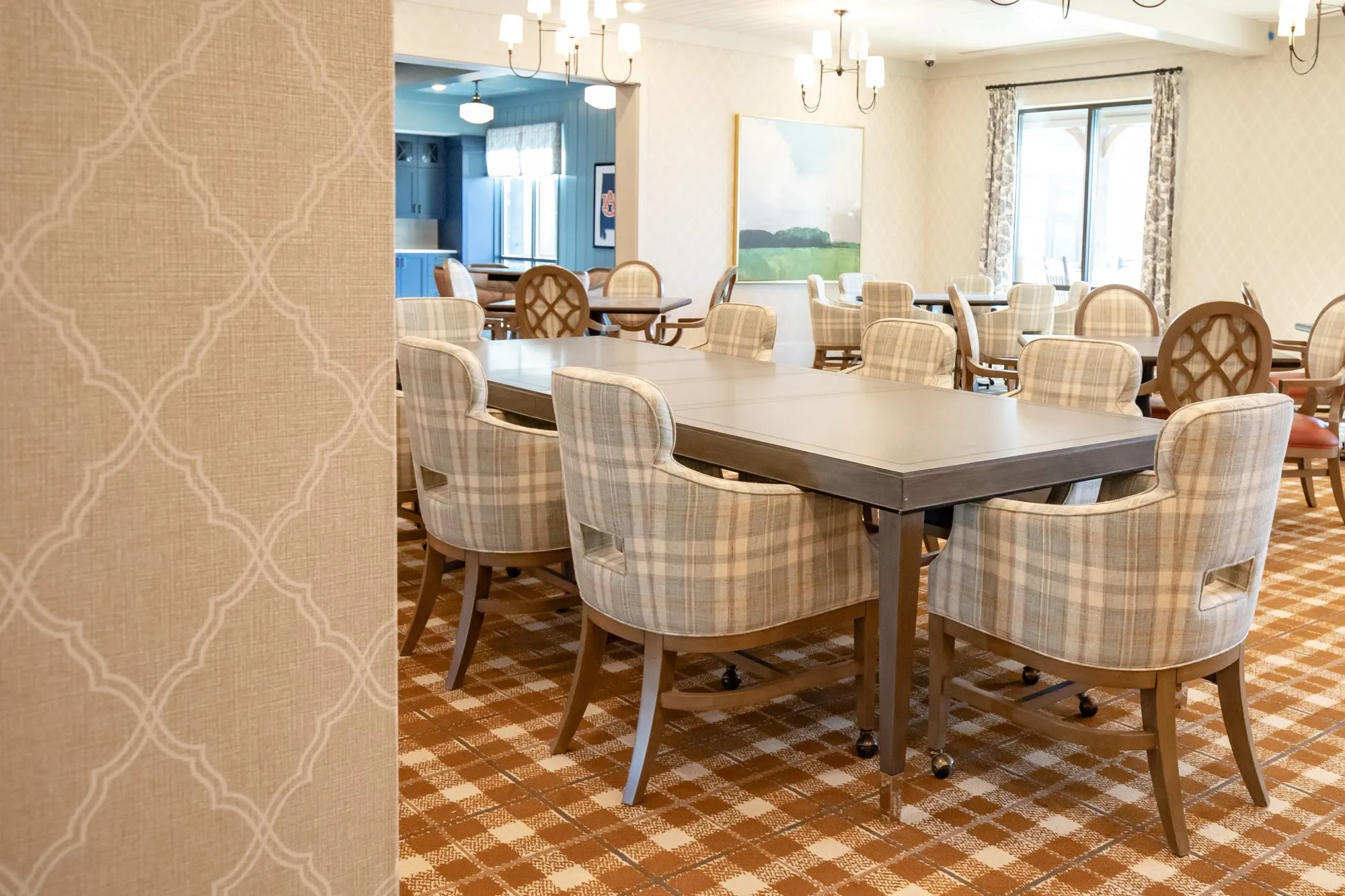 Senior living dining room with a long table of plaid chairs seen past trellis wallcovering, a landscape painting beyond