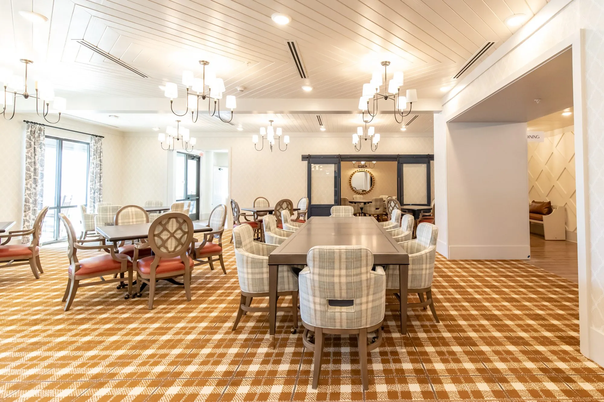 Senior living dining room with plaid chairs at a long table and coral seated chairs under chandeliers
