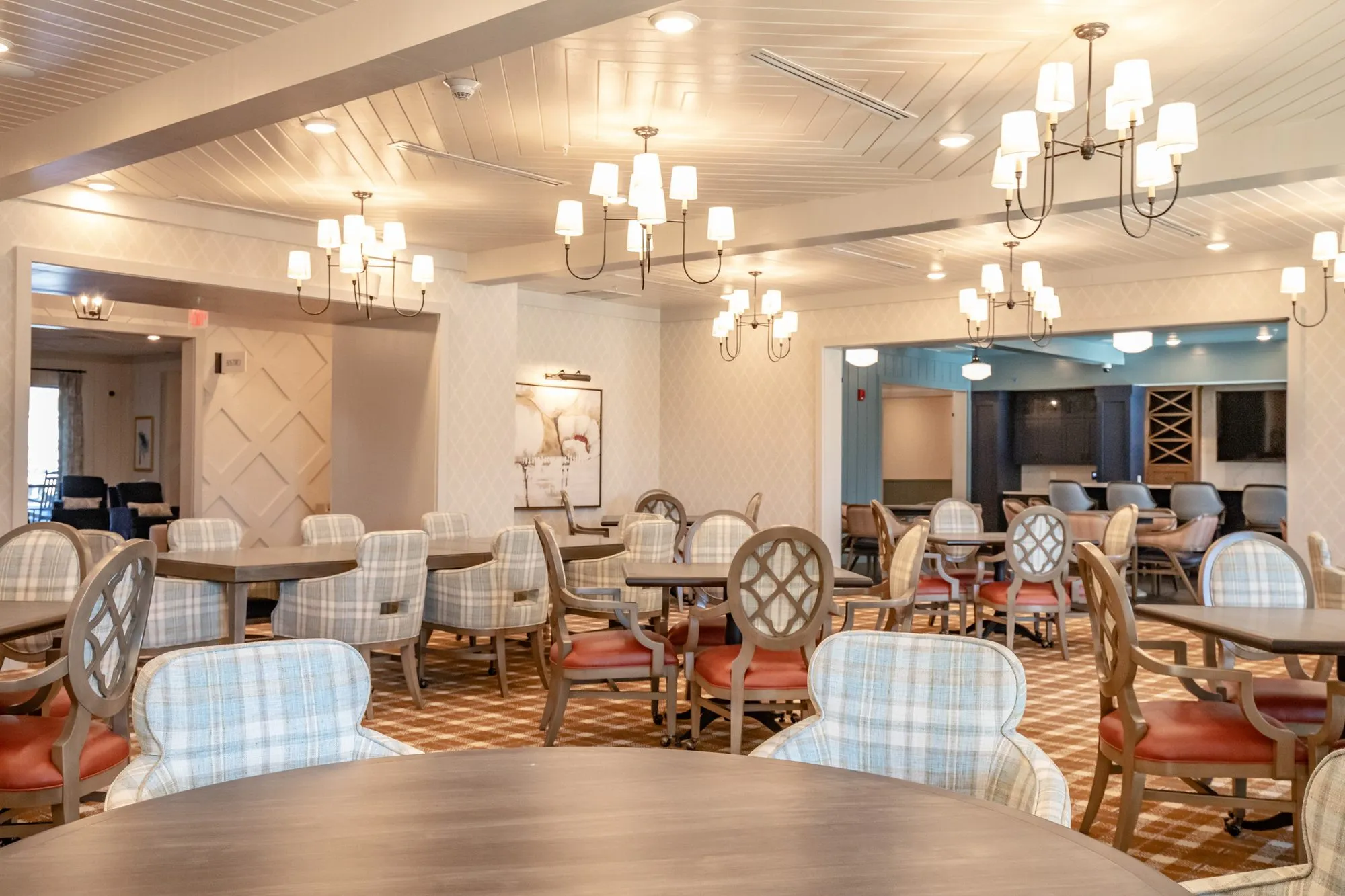 Senior living dining room from a round table, quatrefoil and plaid chairs under chandeliers, the bar beyond