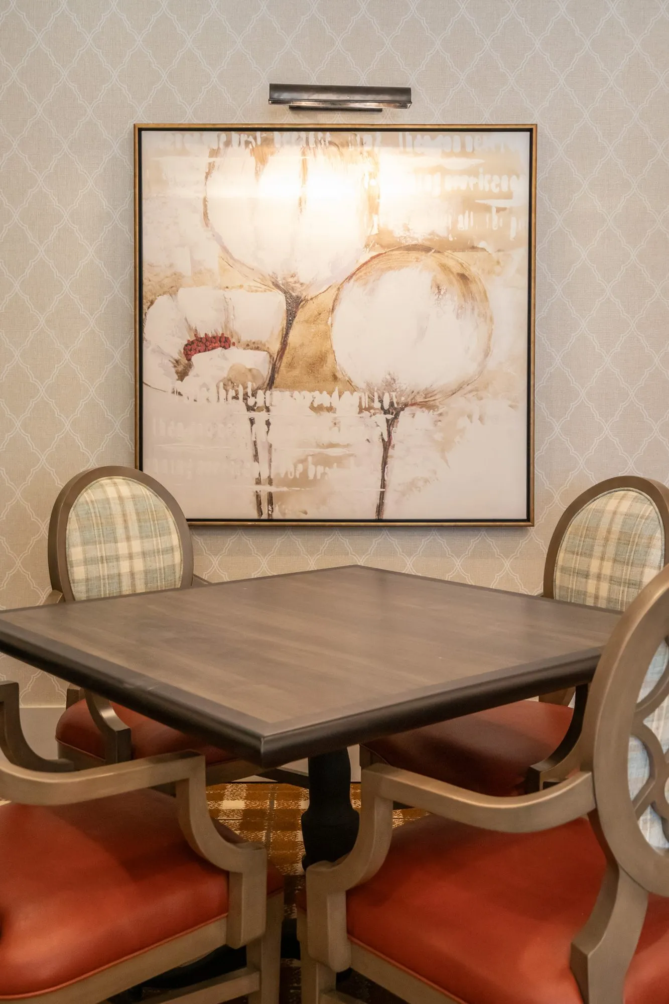 Senior living interior detail: square table with quatrefoil chairs below a cotton boll painting with a picture light
