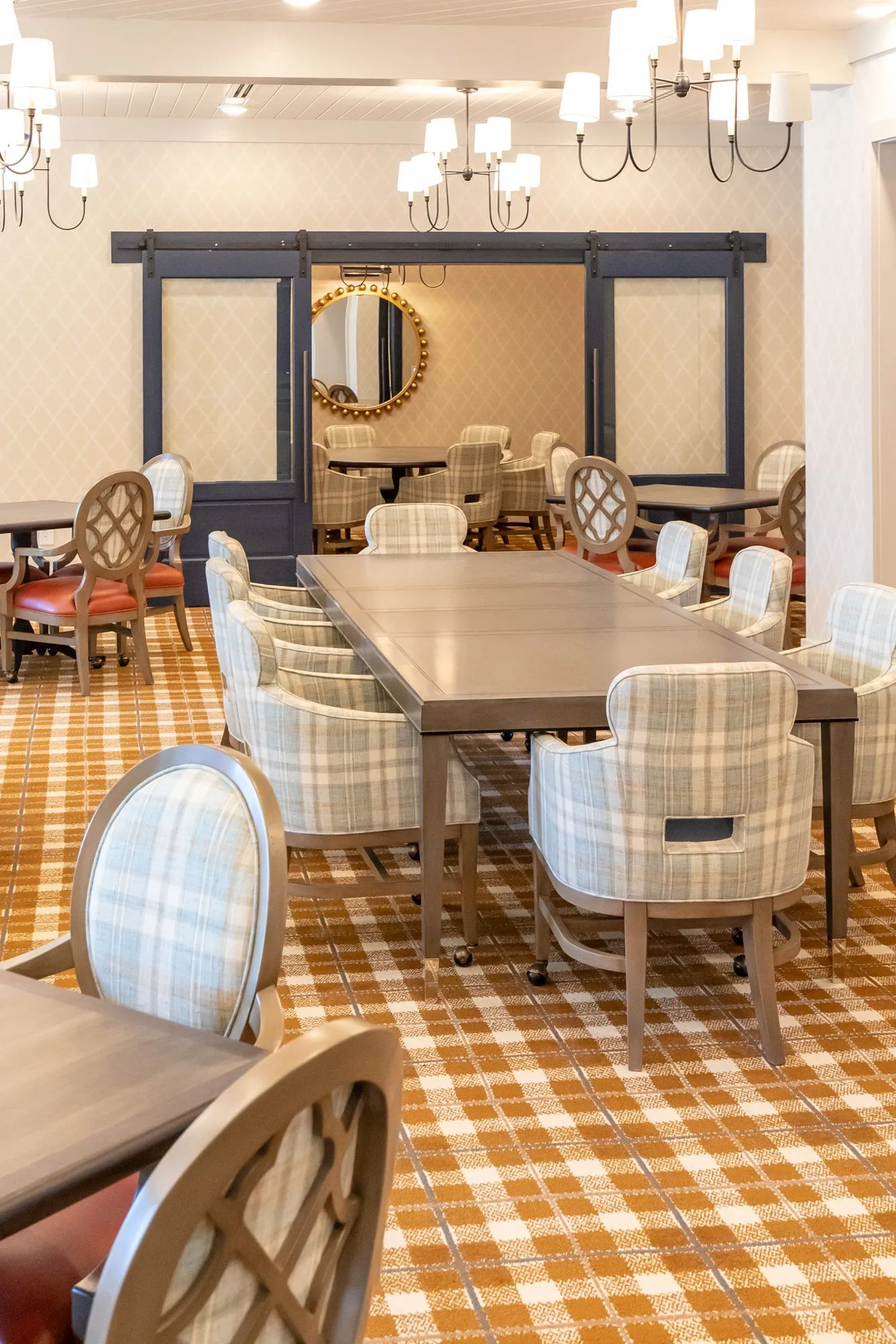 Senior living dining room with a long table of plaid chairs toward barn doors and a gilt beaded mirror