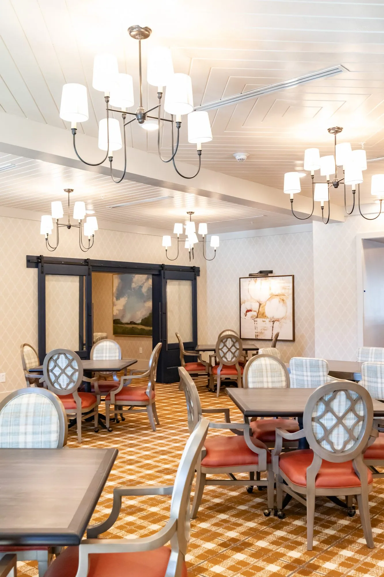 Senior living dining room with coral seated quatrefoil chairs at square tables under candle chandeliers