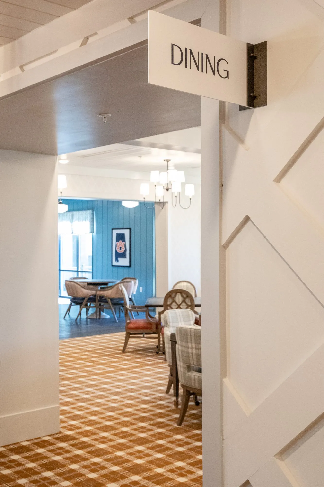 Senior living dining room entrance under a Dining sign, plaid carpet and chairs by blue shiplap beyond