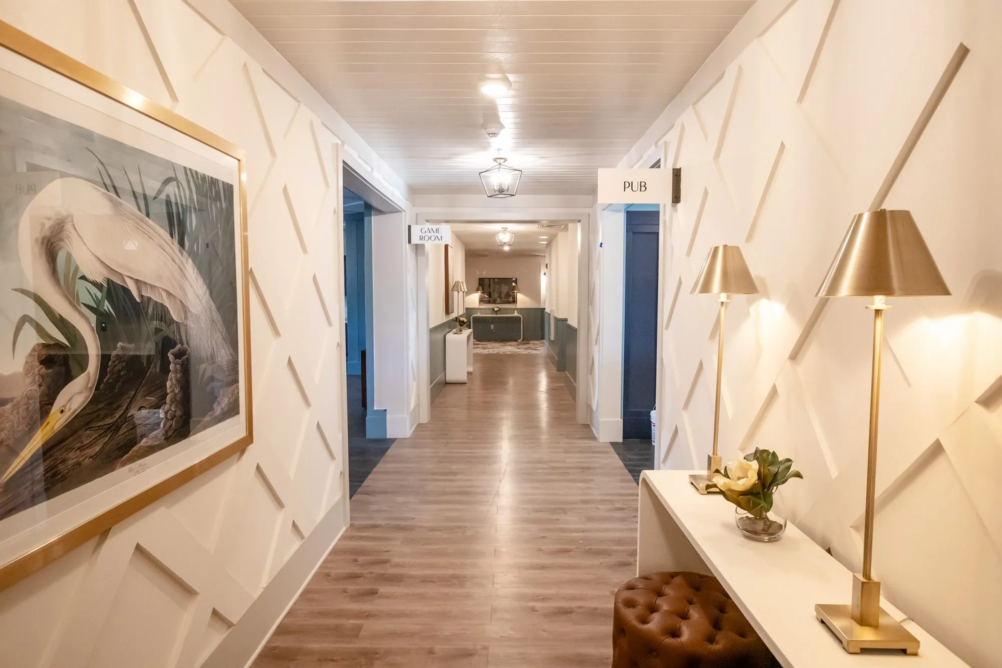 Senior living corridor with an egret print, diamond panelled walls, brass lamps on a console and Pub sign beyond