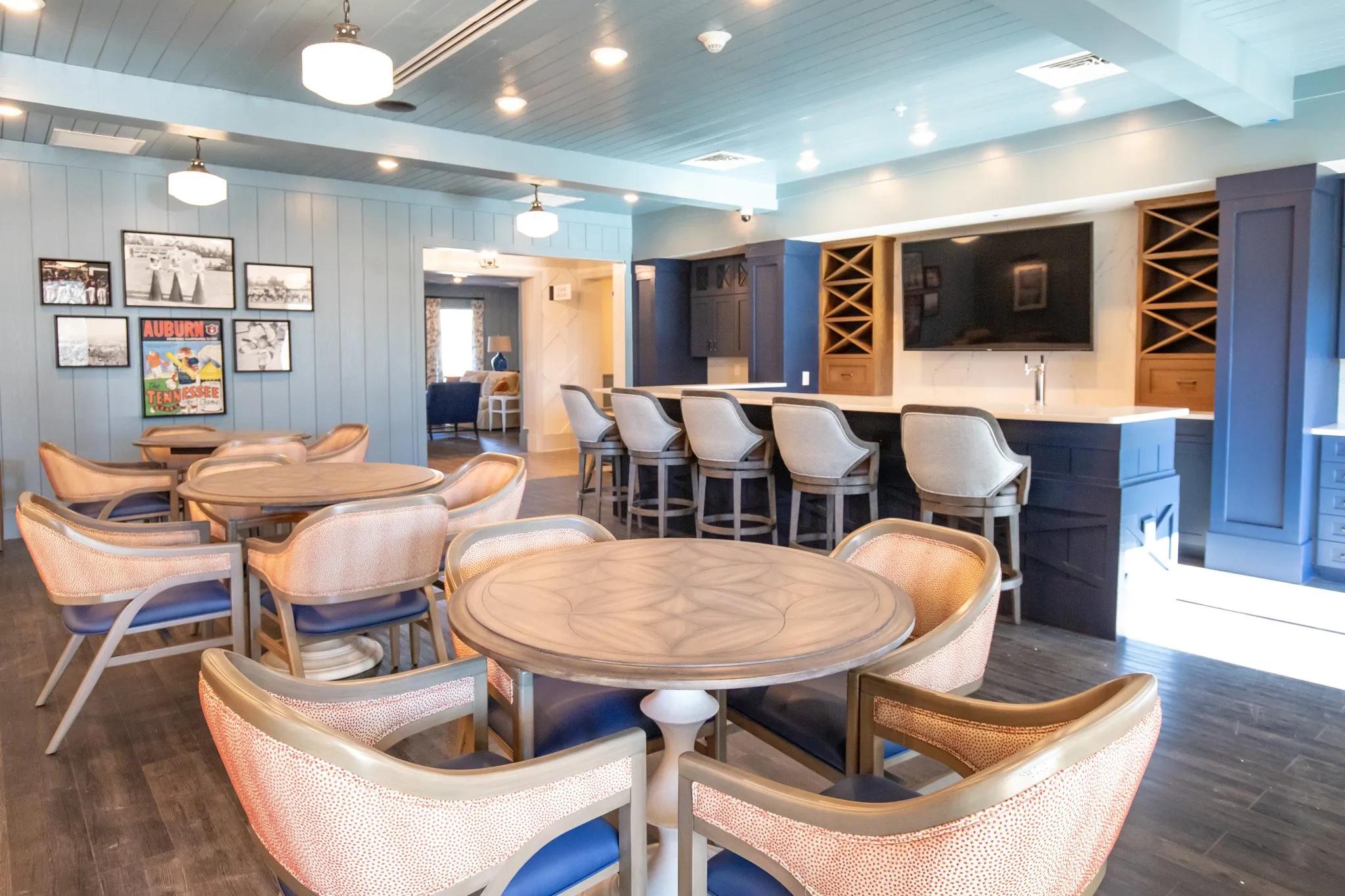 Senior living pub with coral dot chairs at round tables, a bar with wine racks and a TV, black and white sports photos