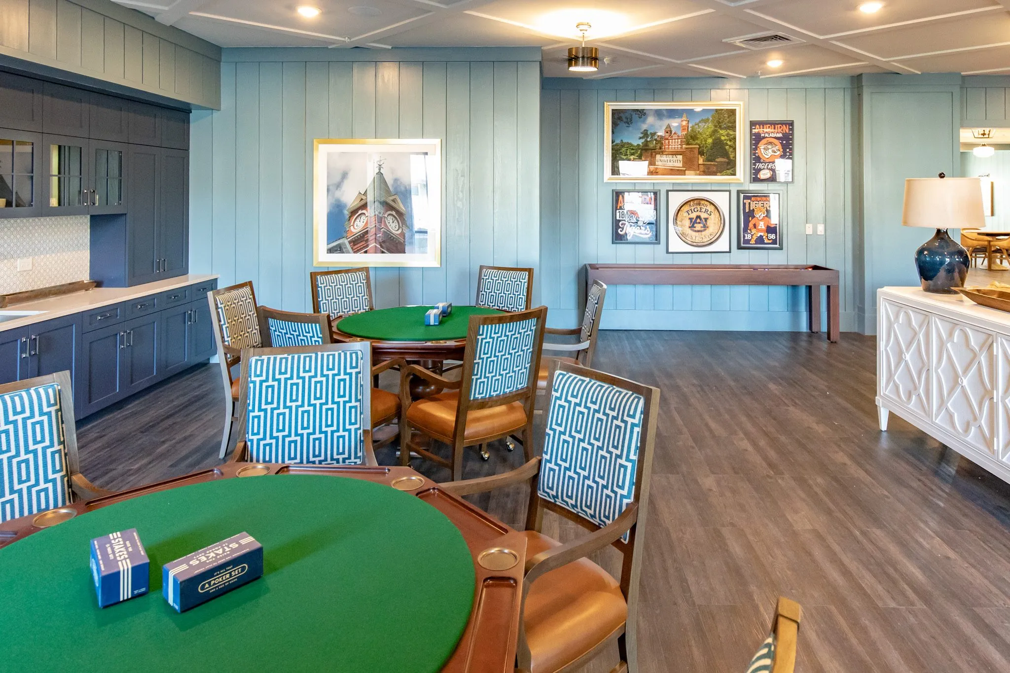 Senior living card room with green felt tables ringed with blue Greek key chairs below framed collegiate prints